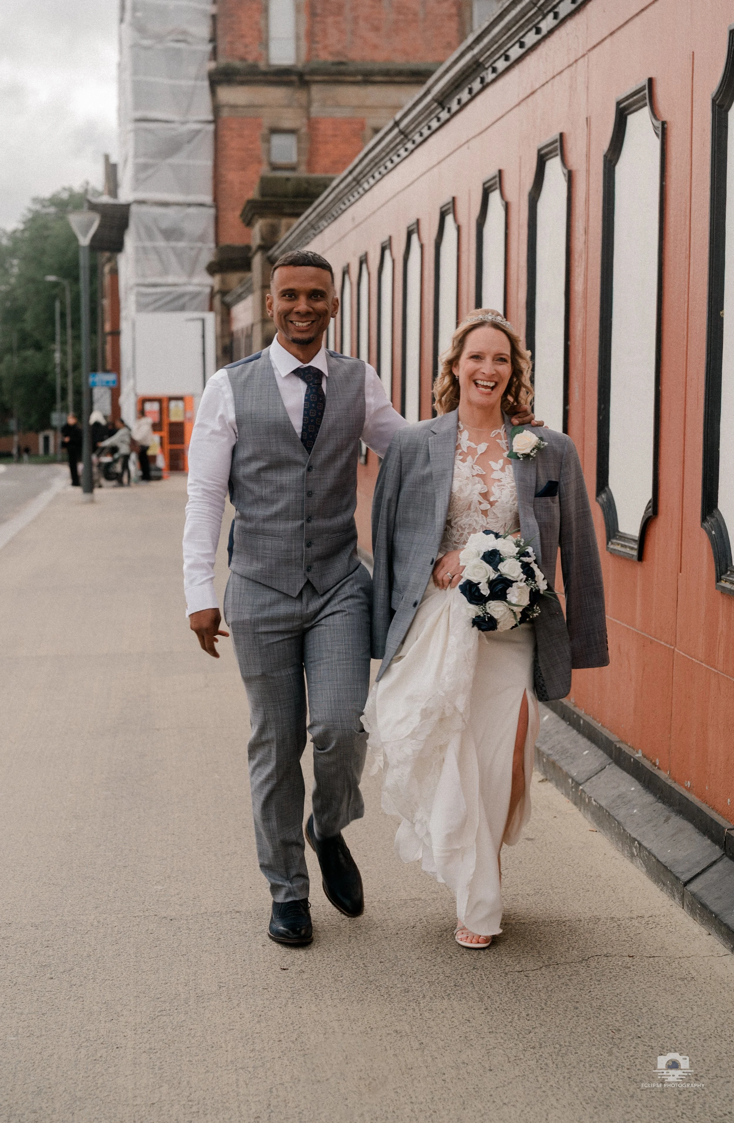 A happy bride and groom walking outside on their wedding day, with the groom in a gray plaid suit and the bride in a white wedding dress holding a bouquet of white and dark flowers, smiling and enjoying their special moment.