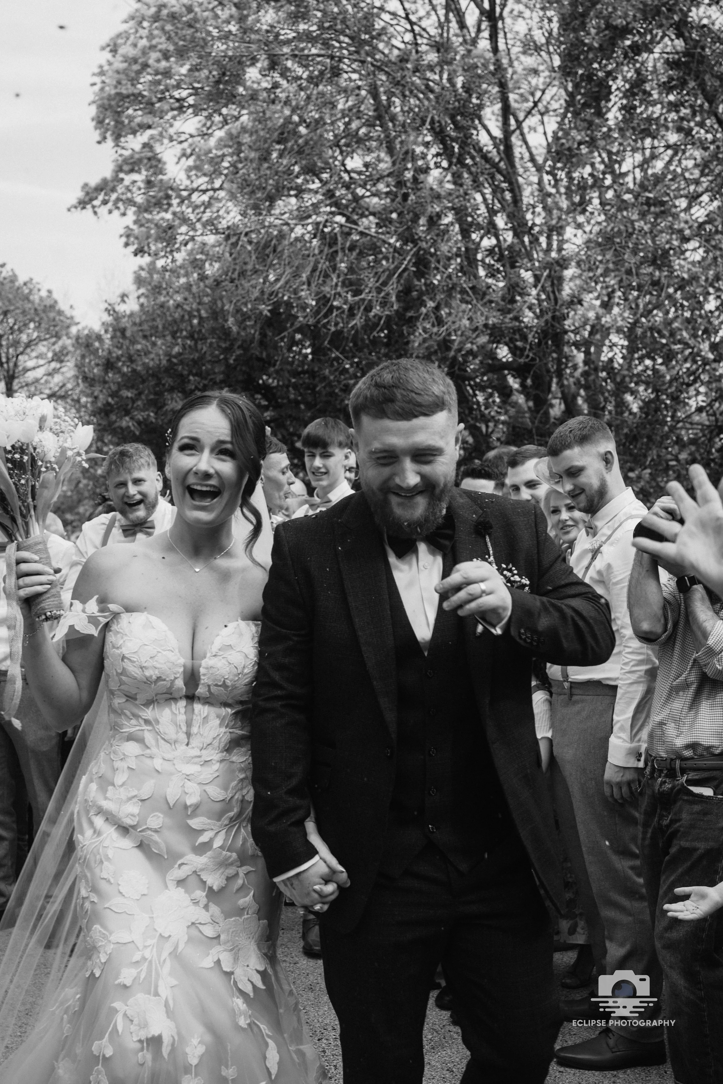 Black and white photo of a newlywed couple outdoors, holding hands and smiling as the groom looks down. The bride wears a strapless wedding dress with floral embroidery and the groom is dressed in a dark suit with a bow tie, surrounded by friends and family.