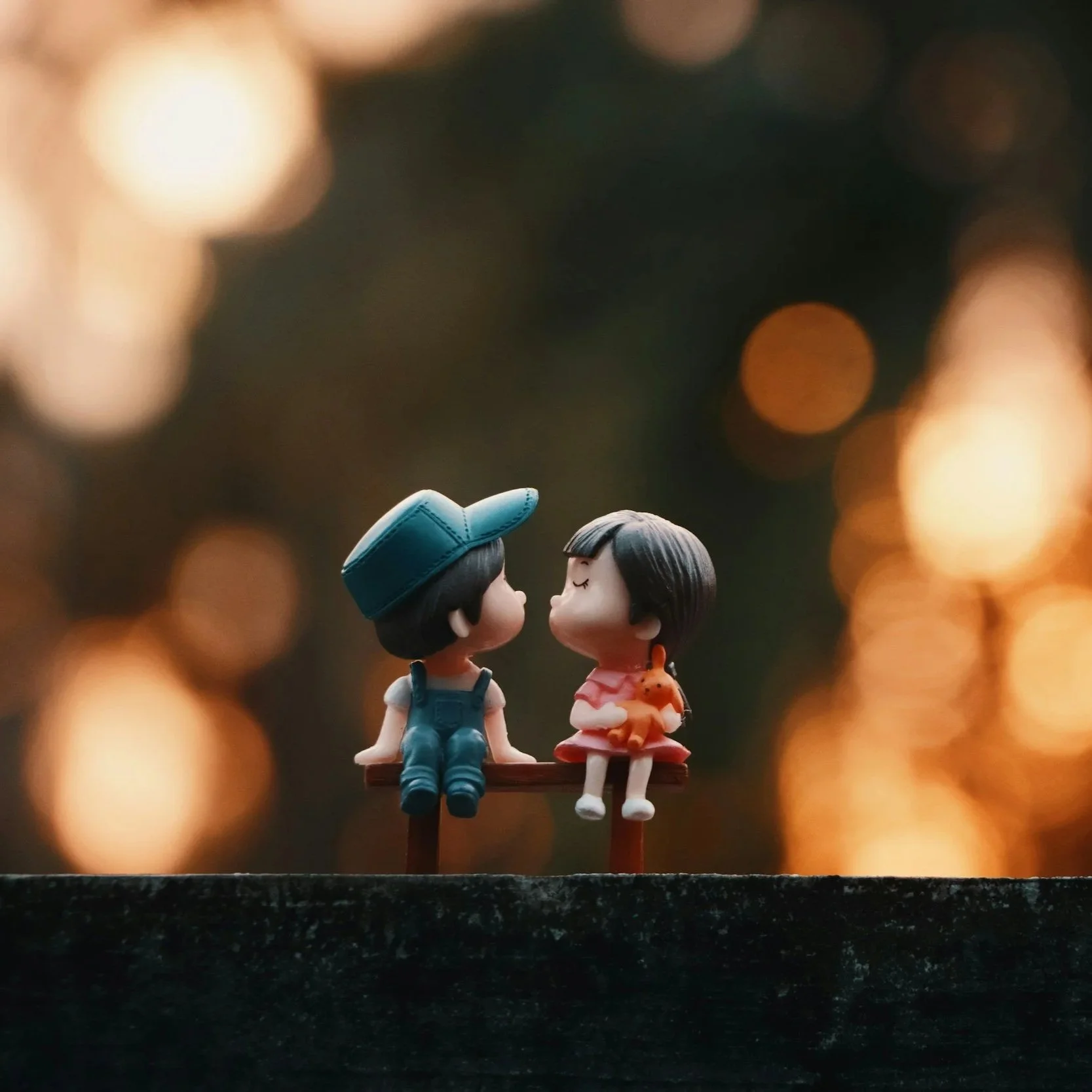 Two small dolls, one wearing a blue hat and the other in a pink dress holding a small orange dog, sitting on a bench against a blurred background with warm bokeh lights.