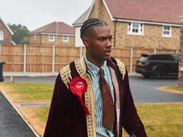 Young man with braided hair wearing traditional graduation attire with a maroon and gold robe and a red sash, standing outdoors near houses and a parked car.
