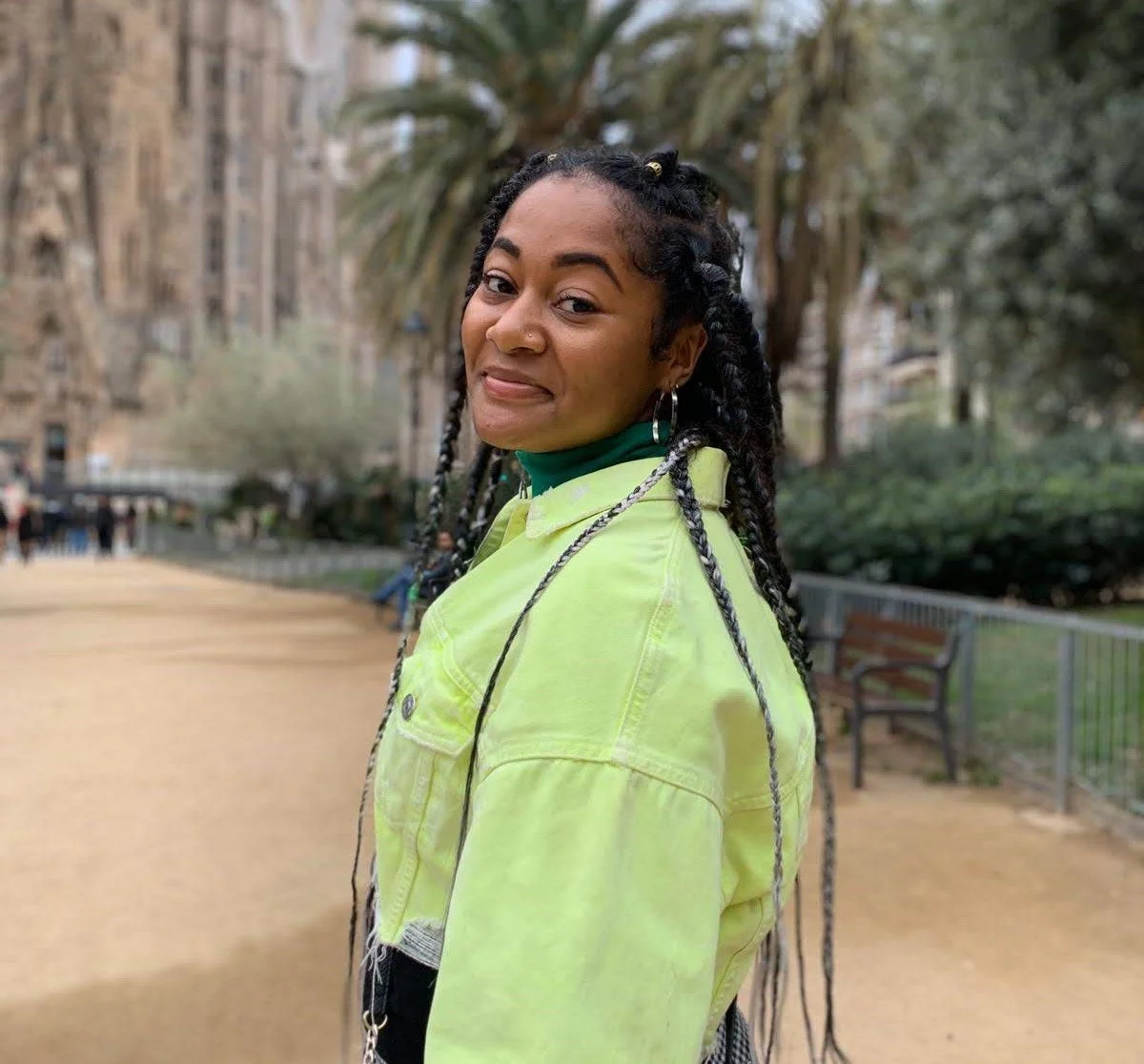 A smiling woman with braids, hoop earrings, wearing a green jacket, outdoors in park with benches, trees, and buildings in background.