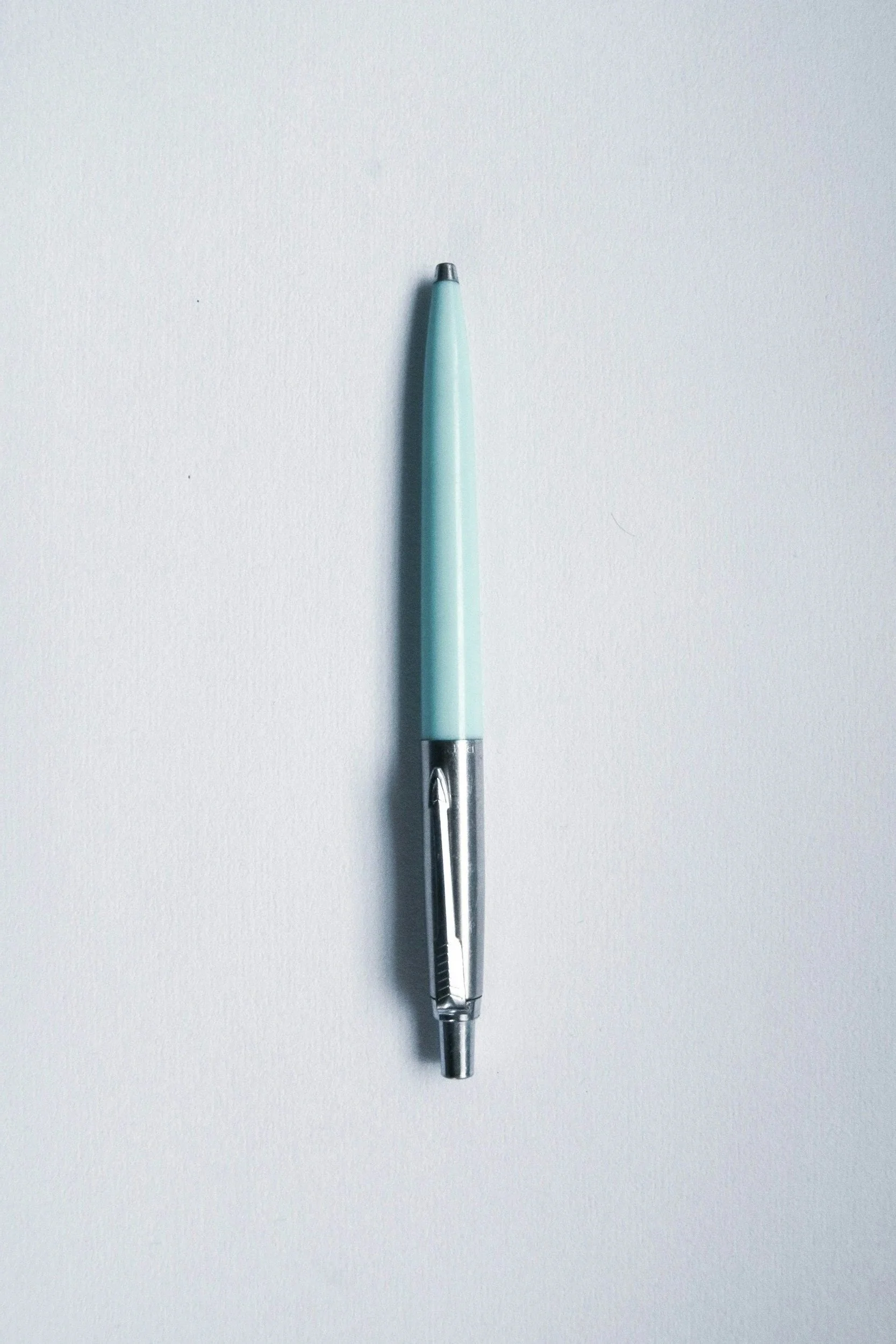 A light blue ballpoint pen with a metallic clip and tip, placed vertically on a blank white surface.