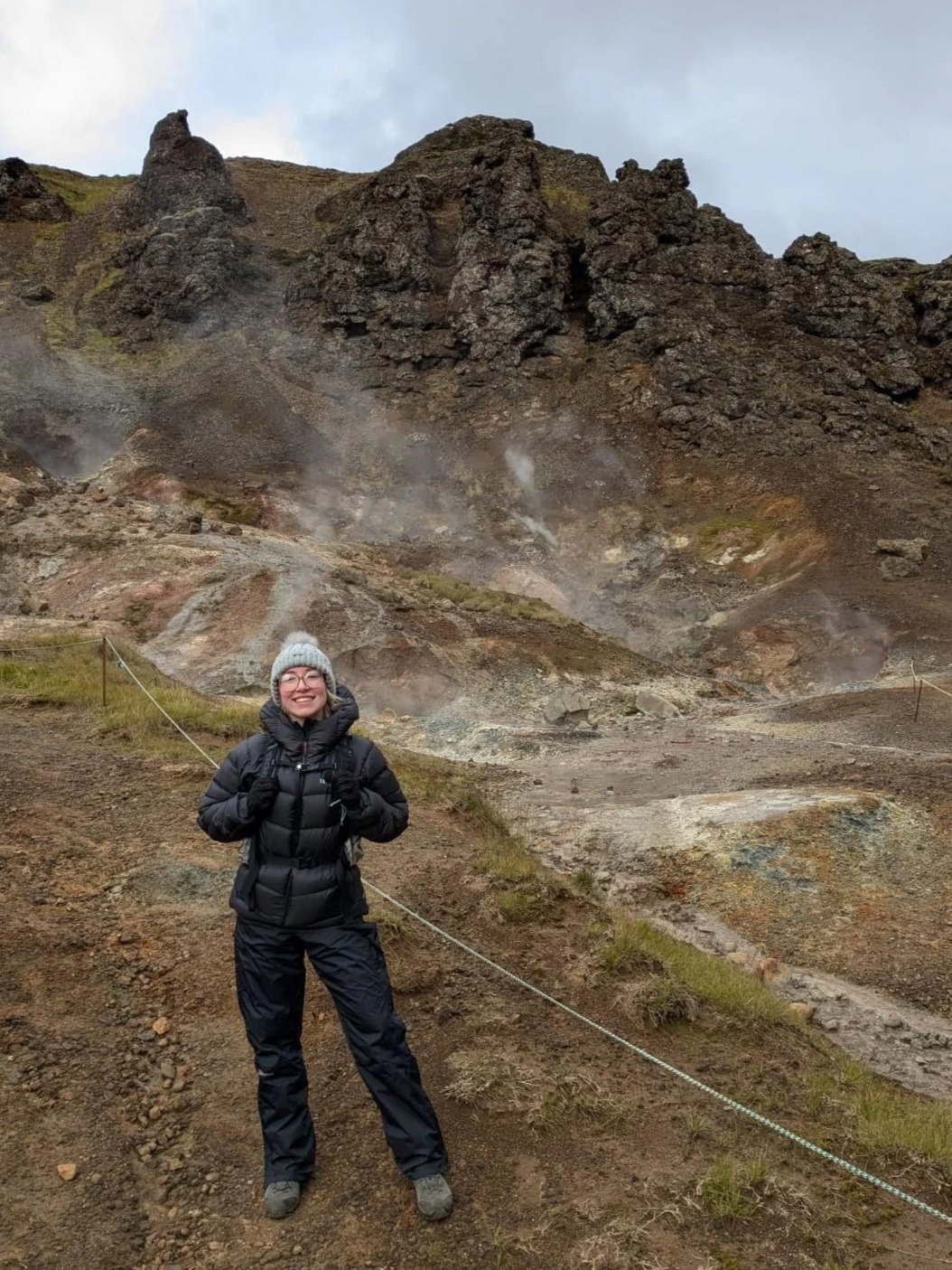 A woman dressed in black outdoor winter clothing and a gray beanie hat is smiling and standing on a rocky trail in front of a volcanic landscape with steam vents and dark volcanic rock formations.