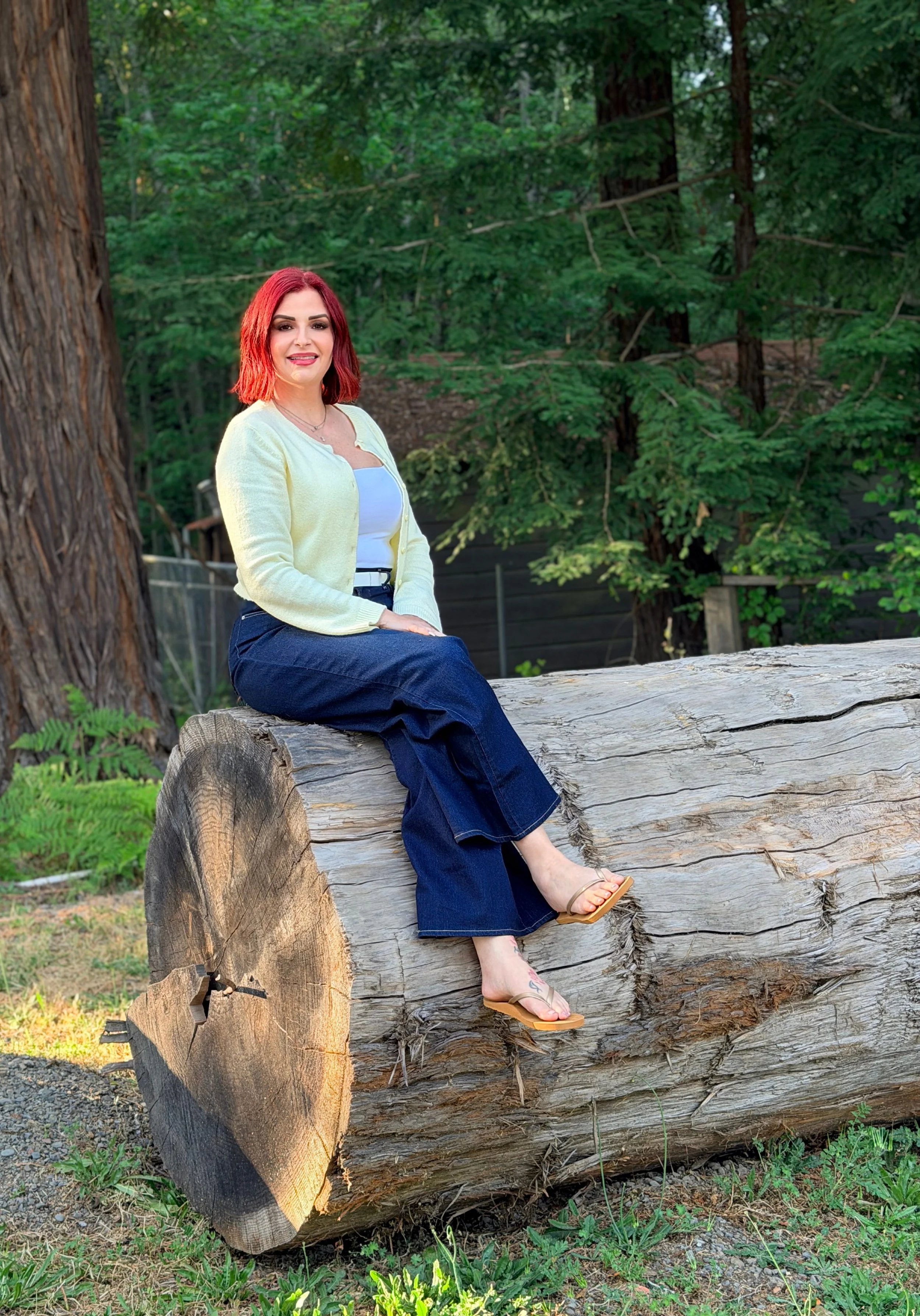 a female with red hair sitting on a log with trees behind her