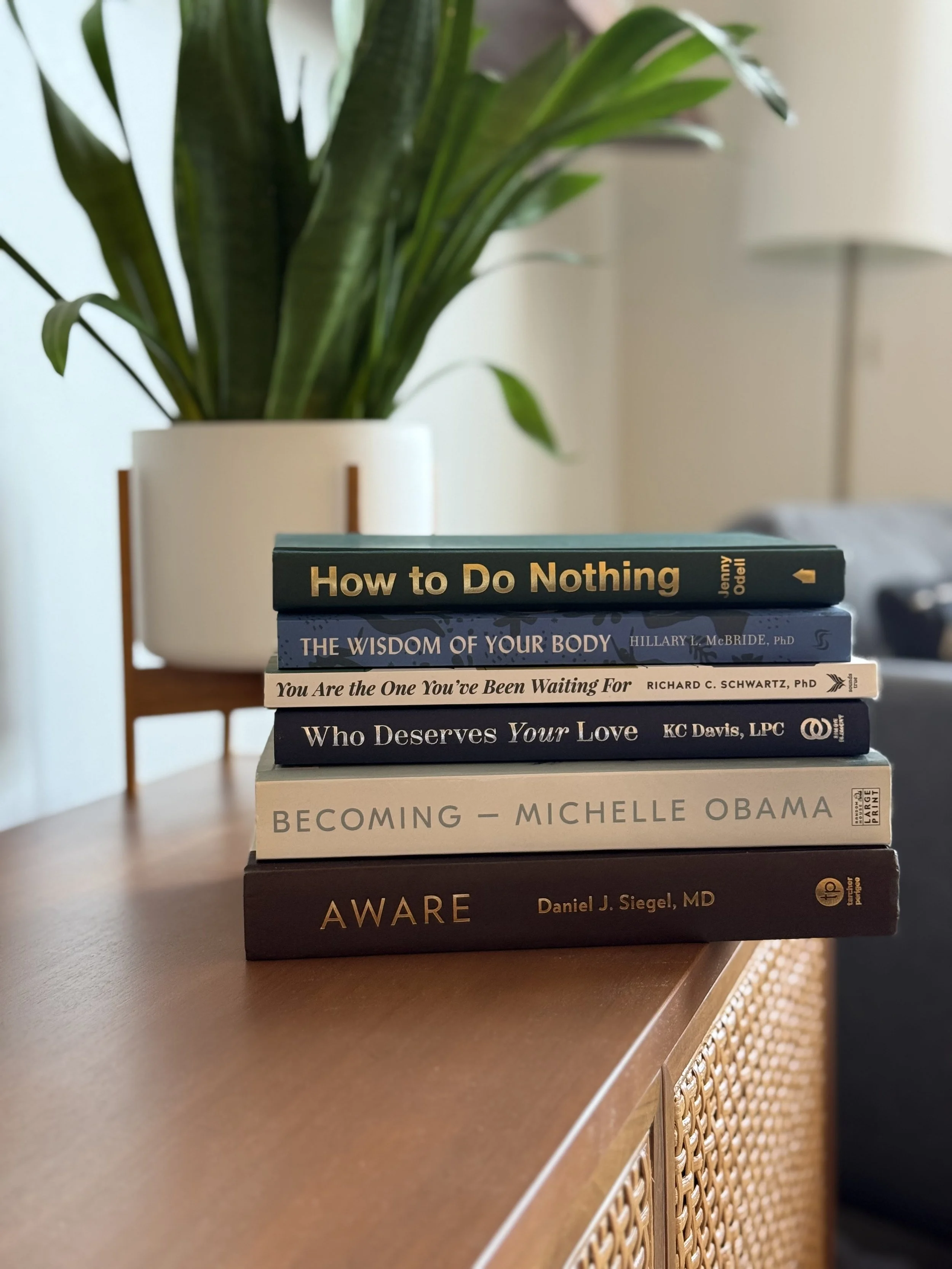 Stack of books on a wooden table in Dr. Heather Stevenson's therapy office, with a plant in the background.