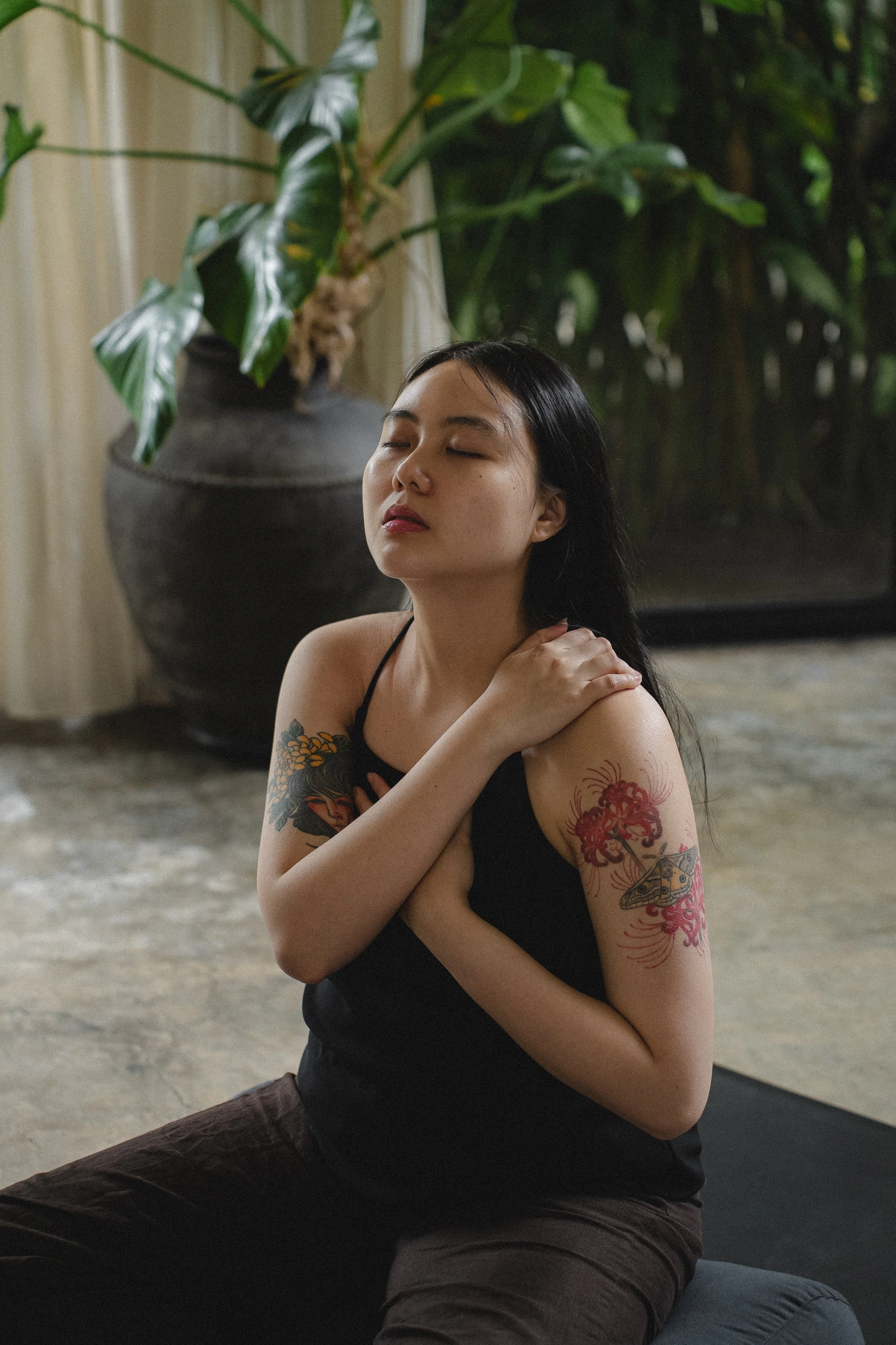 A woman with tattoos on her arm practicing yoga indoors, sitting on a mat with her eyes closed, touching her neck and chest, with large green plants in the background.