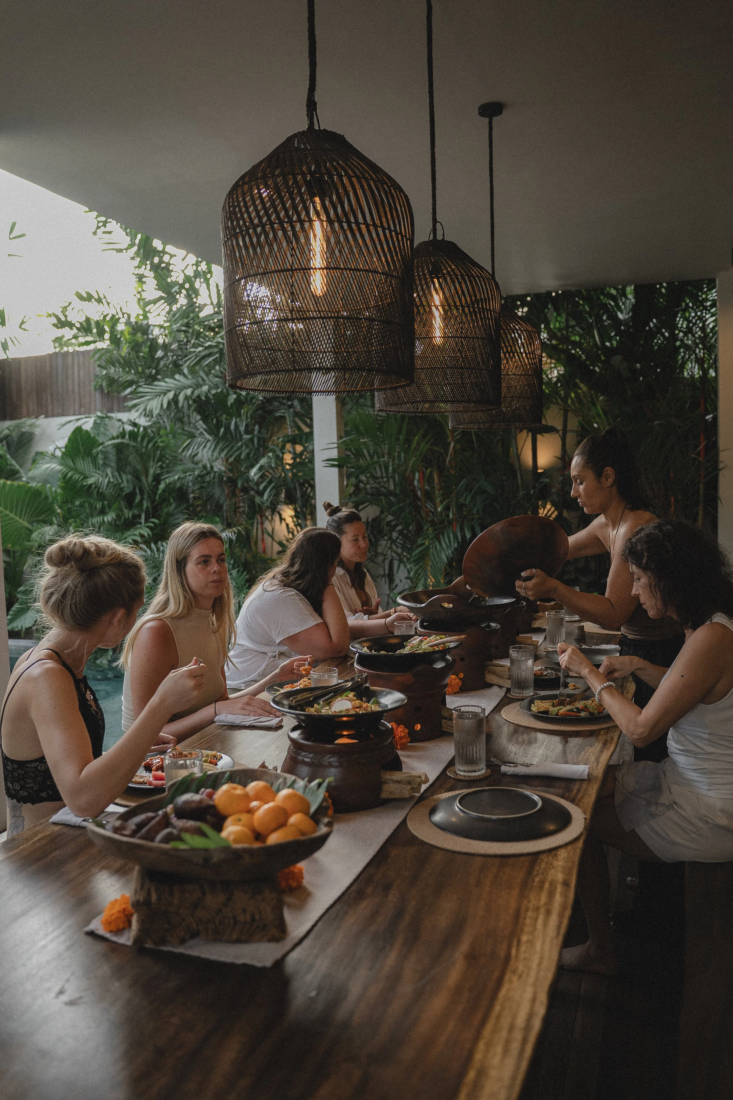 A group of women dining together at a long wooden table in a tropical setting with lush green plants, while a woman pours a drink or food onto a plate. Hanging pendant lights illuminate the scene.