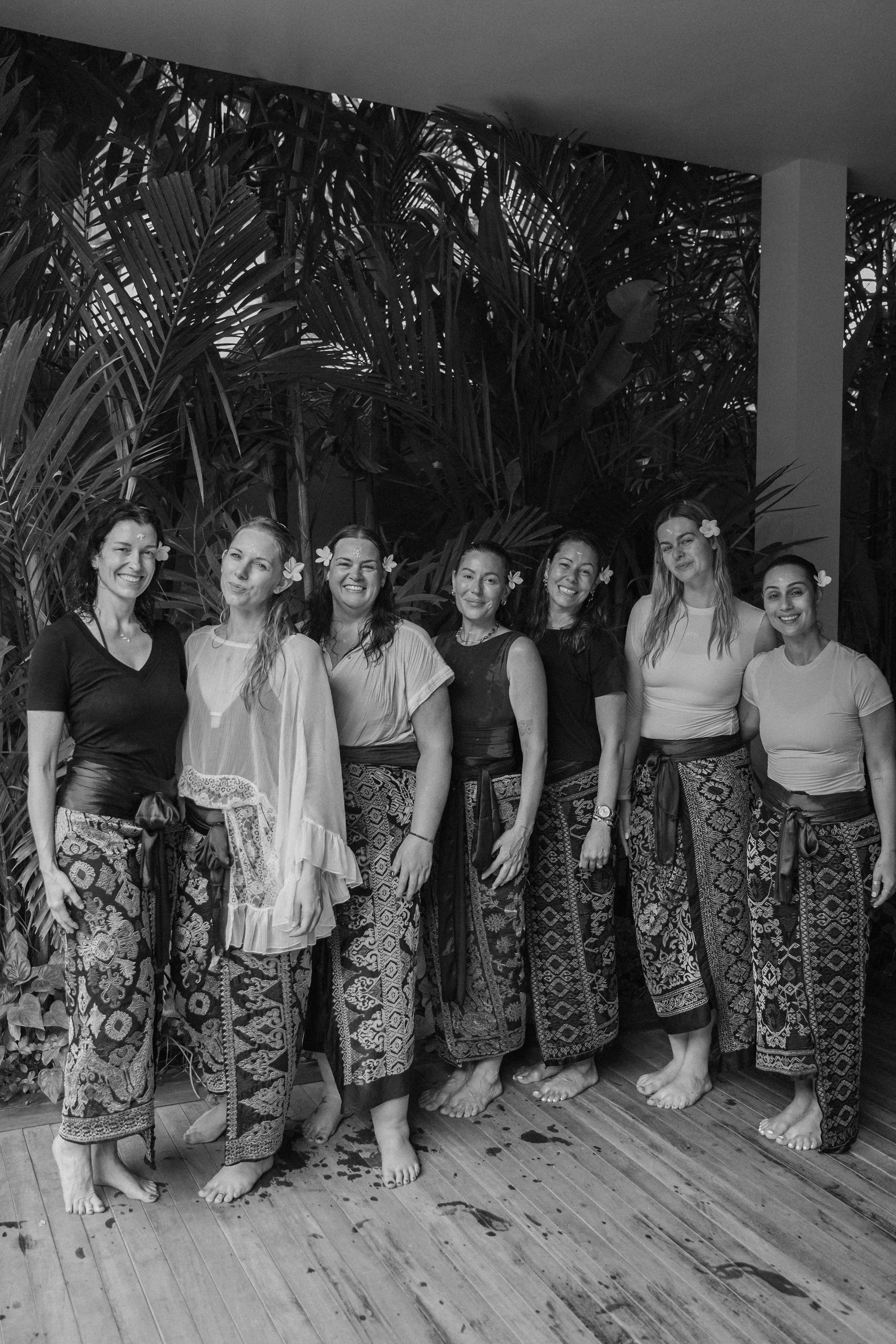 A group of eight women standing together indoors, dressed in traditional patterned wide-leg pants, some with flowers in their hair, on a wooden floor with tropical plants in the background.
