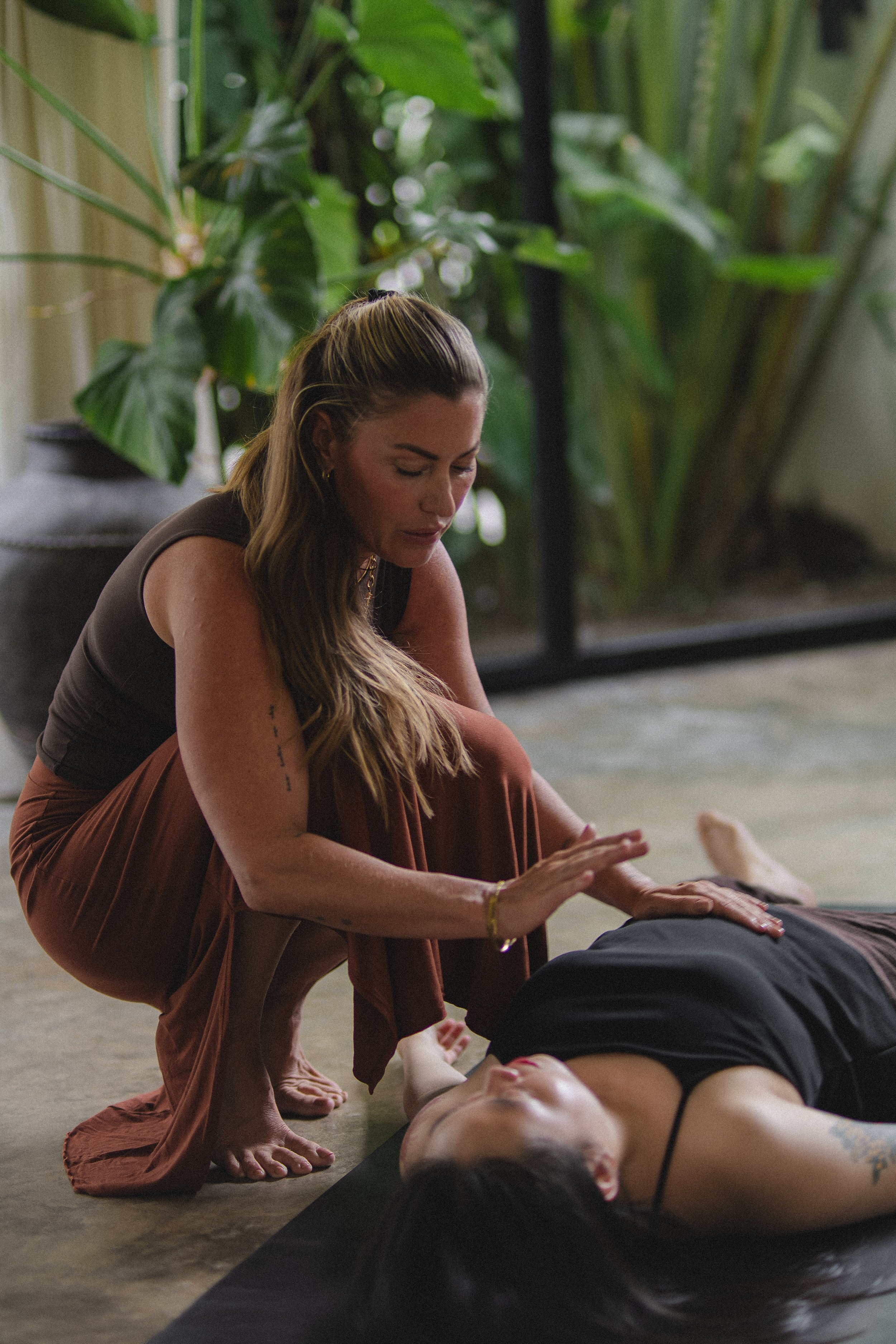 A woman performing CPR on a person lying on a mat on the floor, with green plants and a glass door in the background.