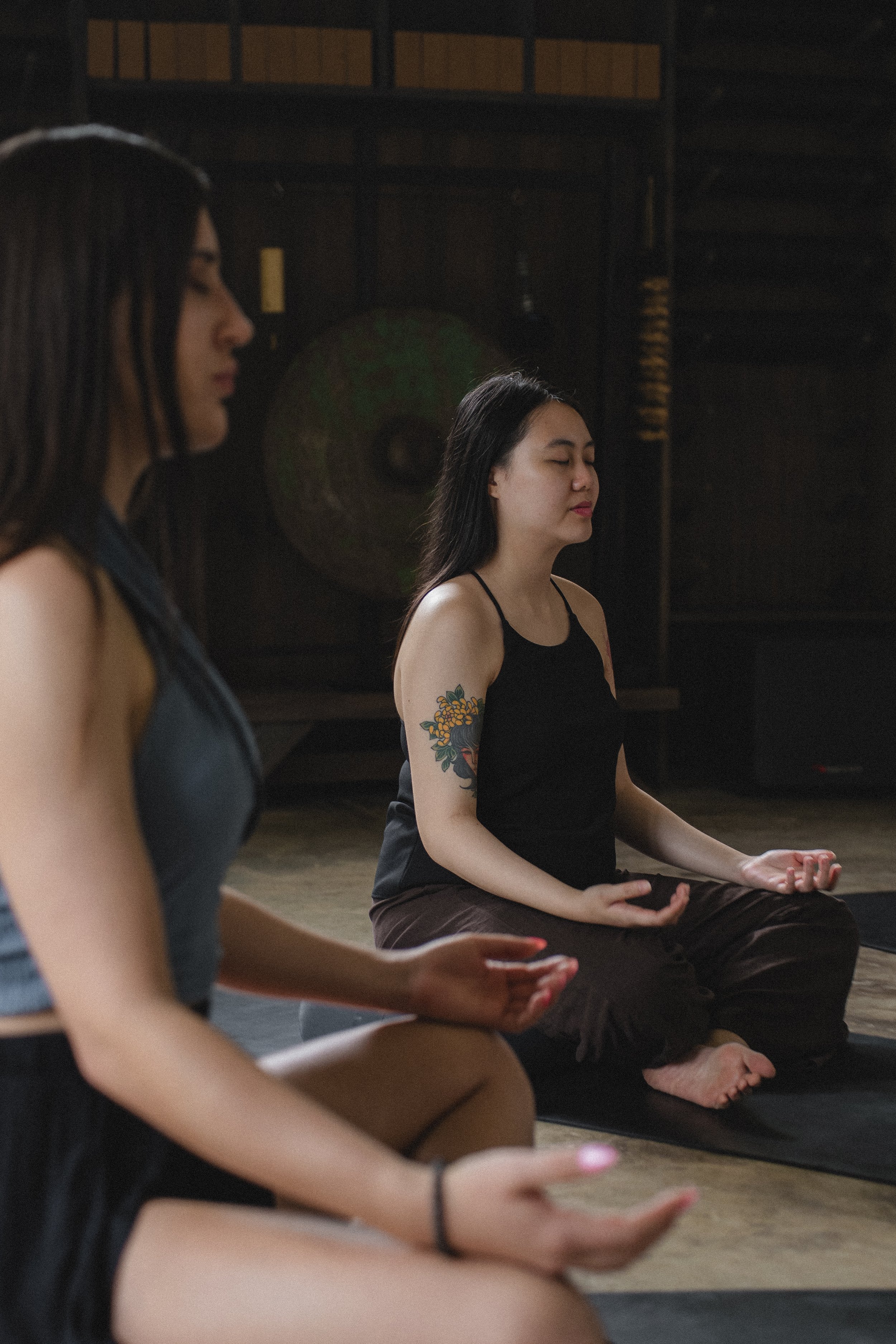 Two women practicing meditation or yoga inside a dimly lit room, sitting cross-legged with eyes closed, one wearing a black tank top and the other in a dark sleeveless top, both on black mats.