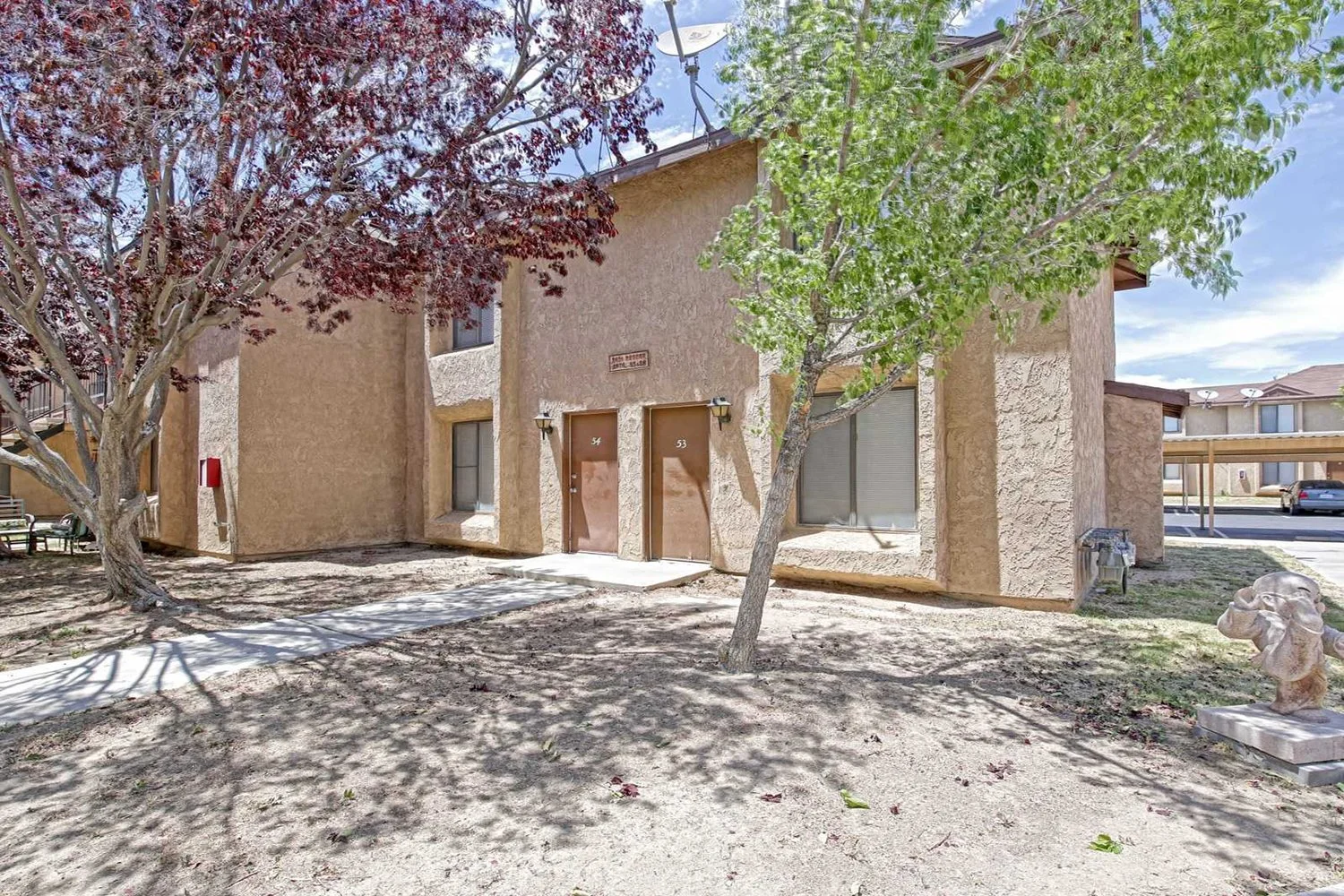 This image shows two front entry doors side-by-side on a two-story tan apartment building surrounded by trees and dirt ground.