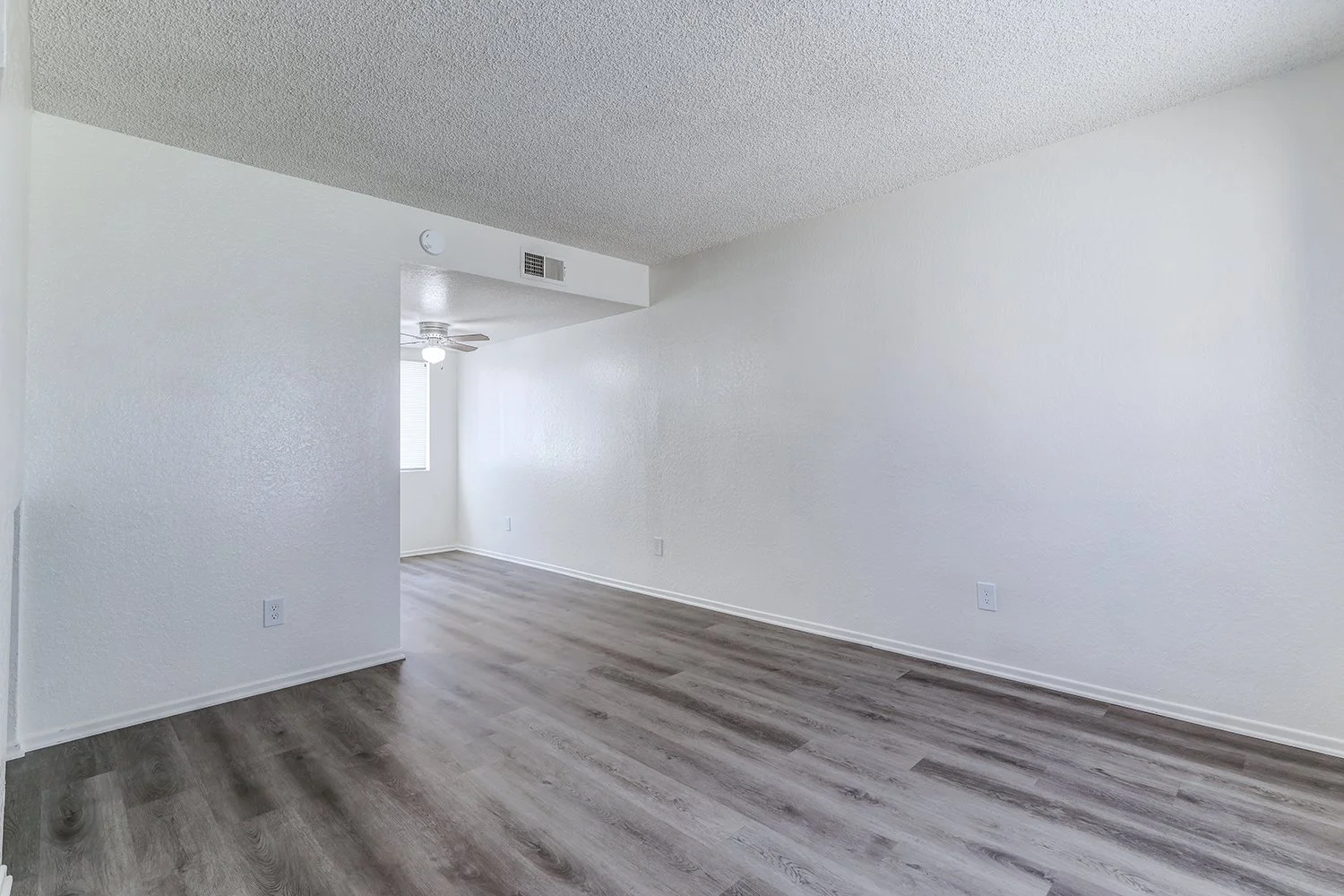 This image shows the interior of an empty apartment living room featuring gray wood-look flooring and plain white walls.