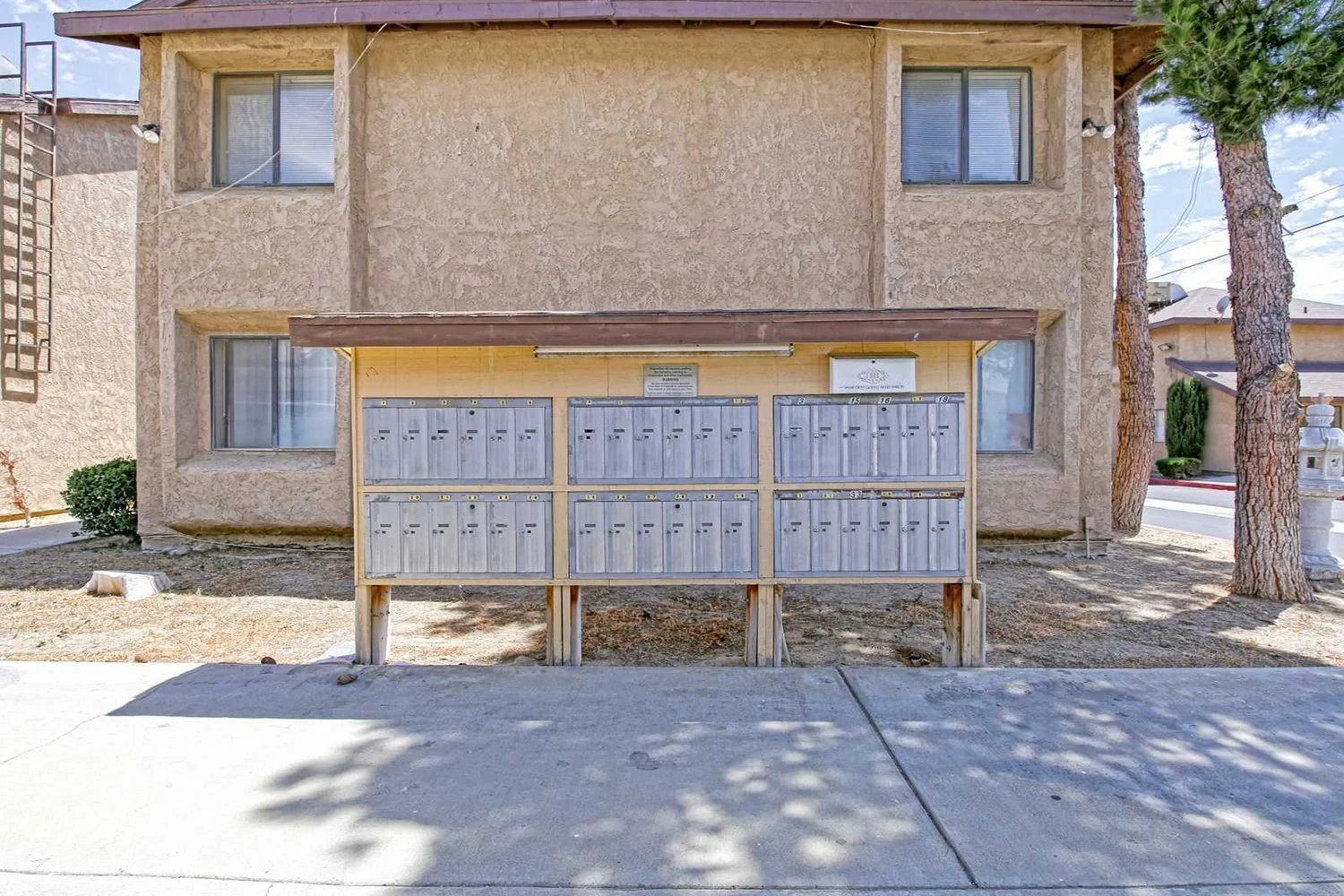 This image shows an outdoor mail with multiple gray metal lockboxes located along a concrete sidewalk in front of a two-story tan apartment building.