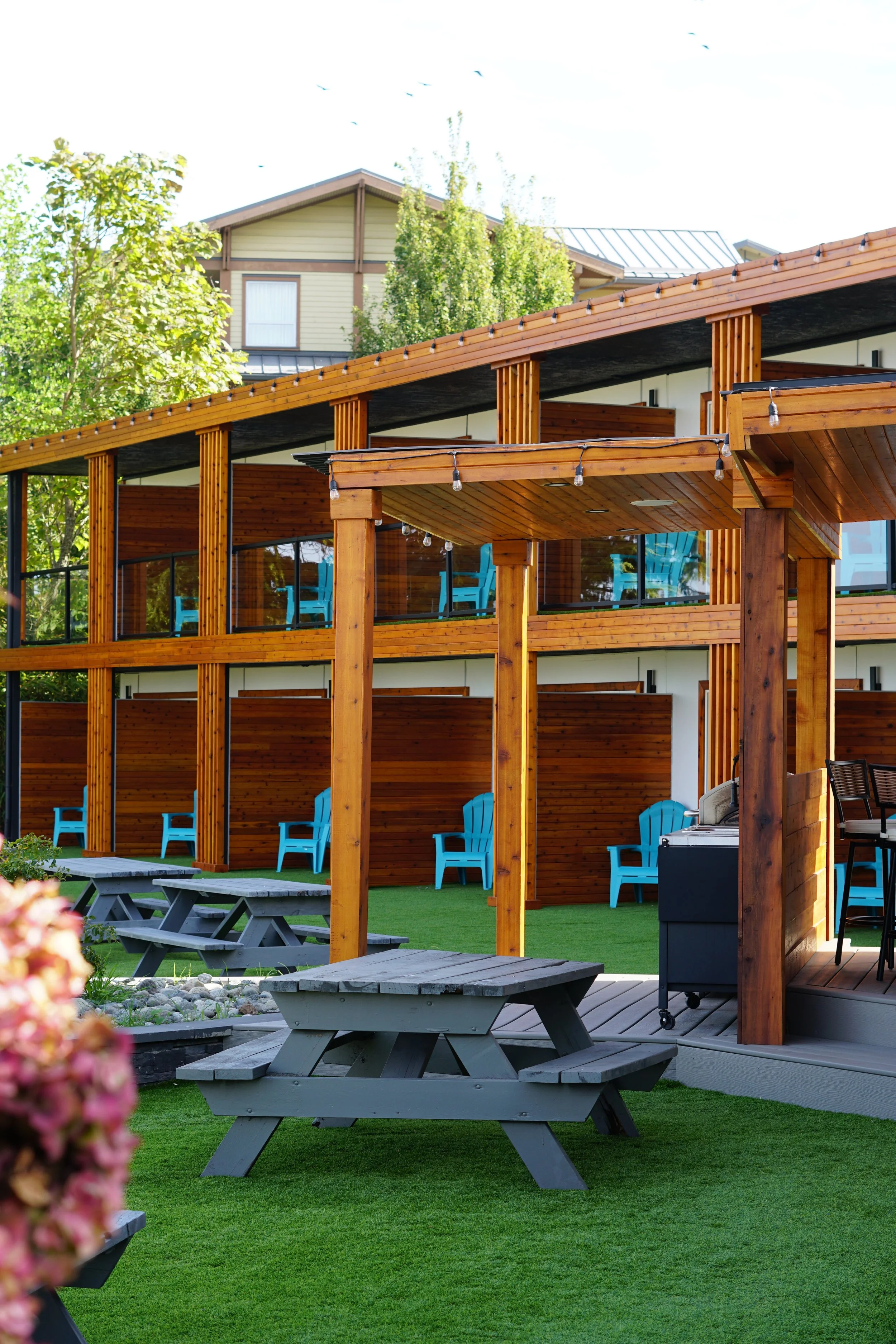 Outdoor patio area with wooden seating and picnic tables, Adirondack chairs, and string lights, adjacent to a modern multi-story building with large glass windows.