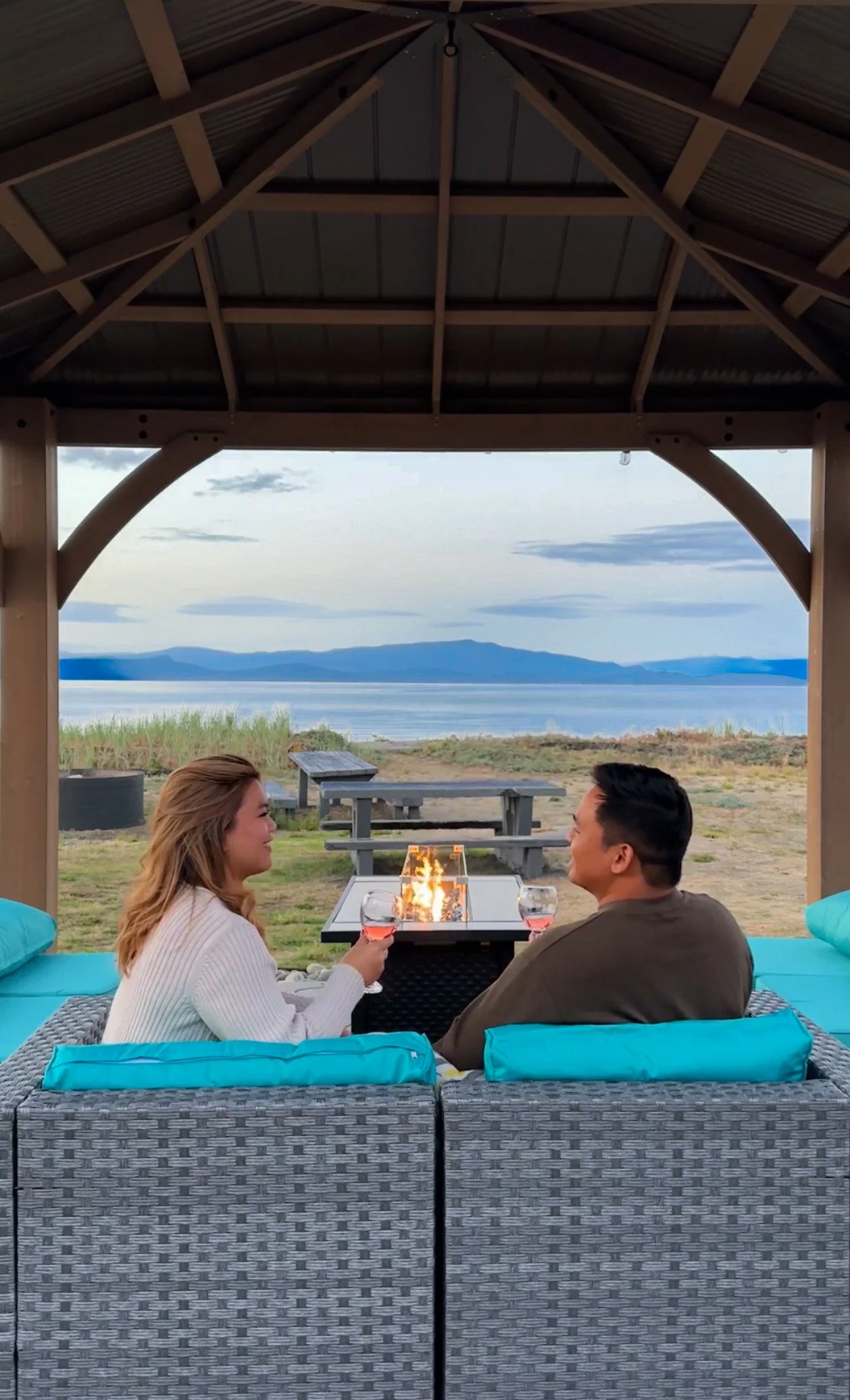A couple sitting on a wicker sofa around a fire pit, holding glasses with pink liquid, under a wooden pavilion overlooking a lake with mountains in the distance.