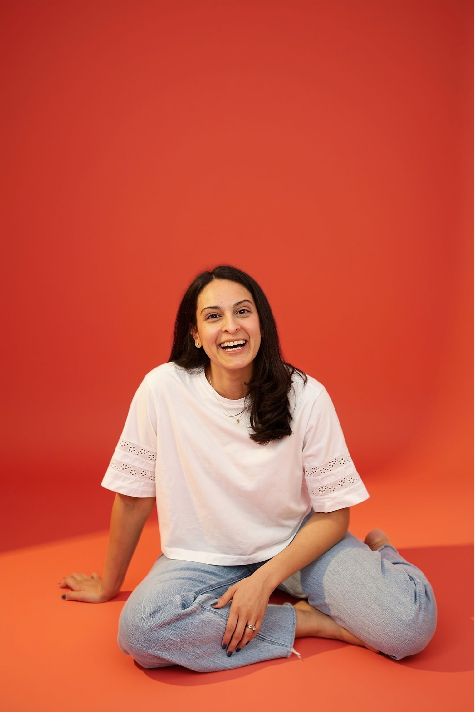 A photo of Jeanine, a licensed counselor, sitting on the floor against an orange background. She wears a white top and light blue jeans with ripped knees.