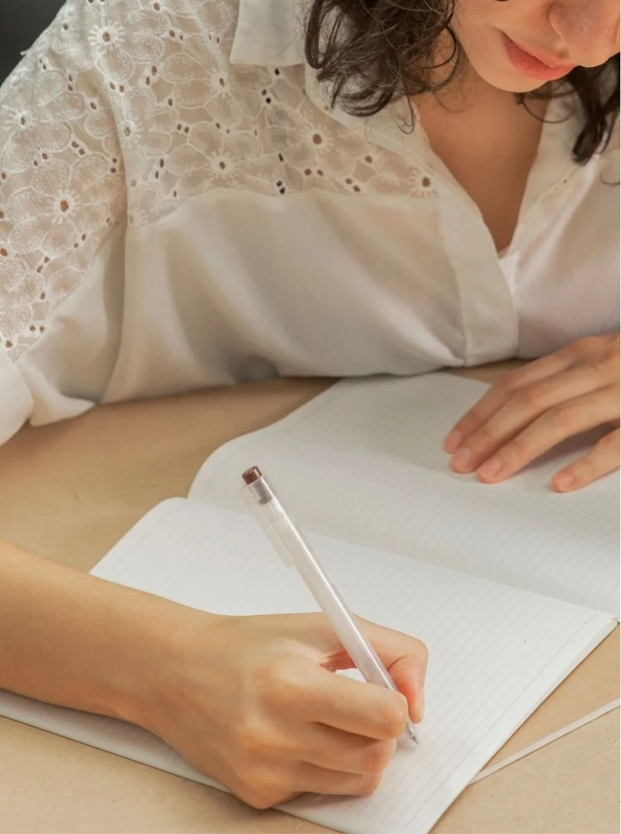 A woman with curly hair wearing a white blouse with lace detailing, sitting at a desk and writing in a therapy notebook.
