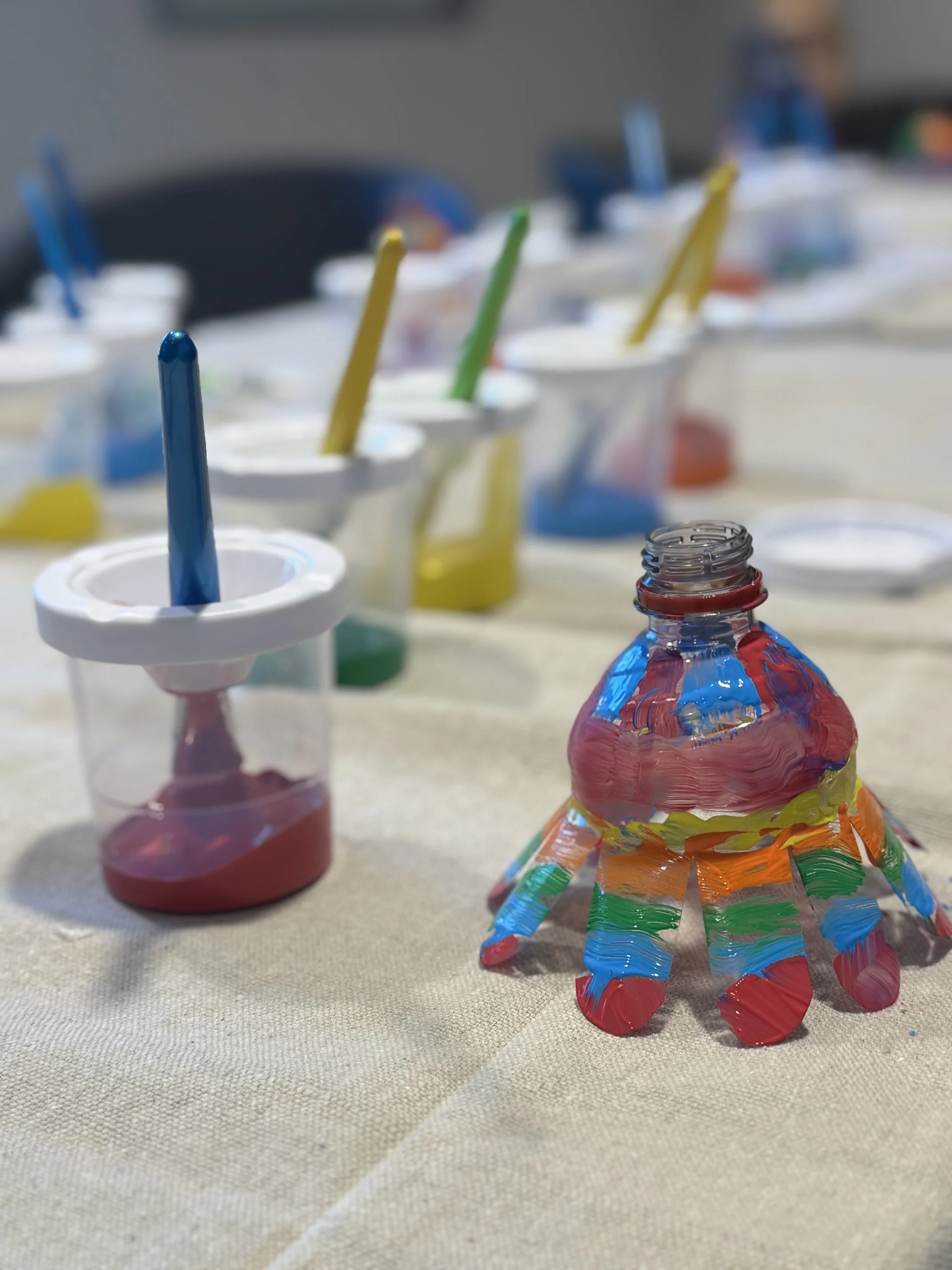 A small, colorful, hand-painted craft resembling a miniature animal or character with multiple legs, made of a bottle or similar material, is placed on a beige fabric surface. In the background, there are small containers of paint with brushes in different colors.