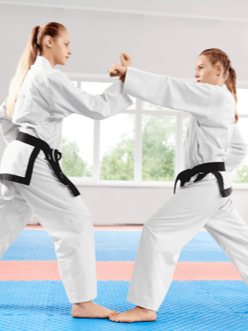 Two young girls practicing karate in a dojo, wearing karate uniforms with black belts, engaged in a sparring match.
