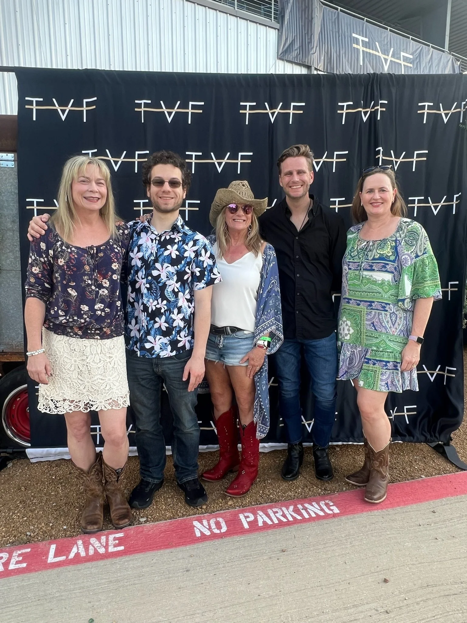 Three people in martial arts uniforms standing together inside a pavilion with trees outside. The man on the left and the woman on the right wear black belts, and the young woman in the middle has a black belt with colorful patches. They are smiling.