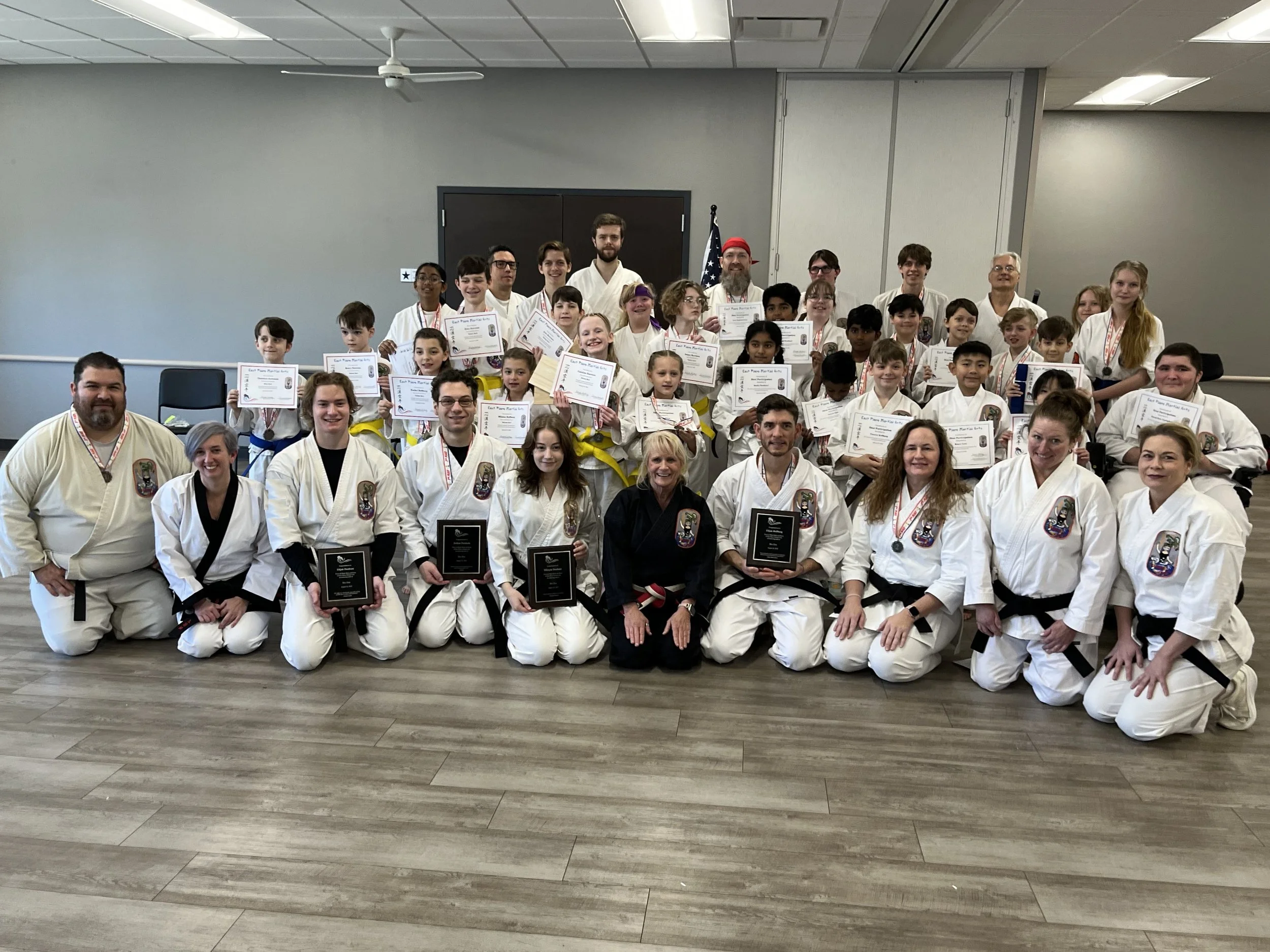 Group of martial artists in white uniforms, some holding gifts and medals, posing indoors on a green mat.