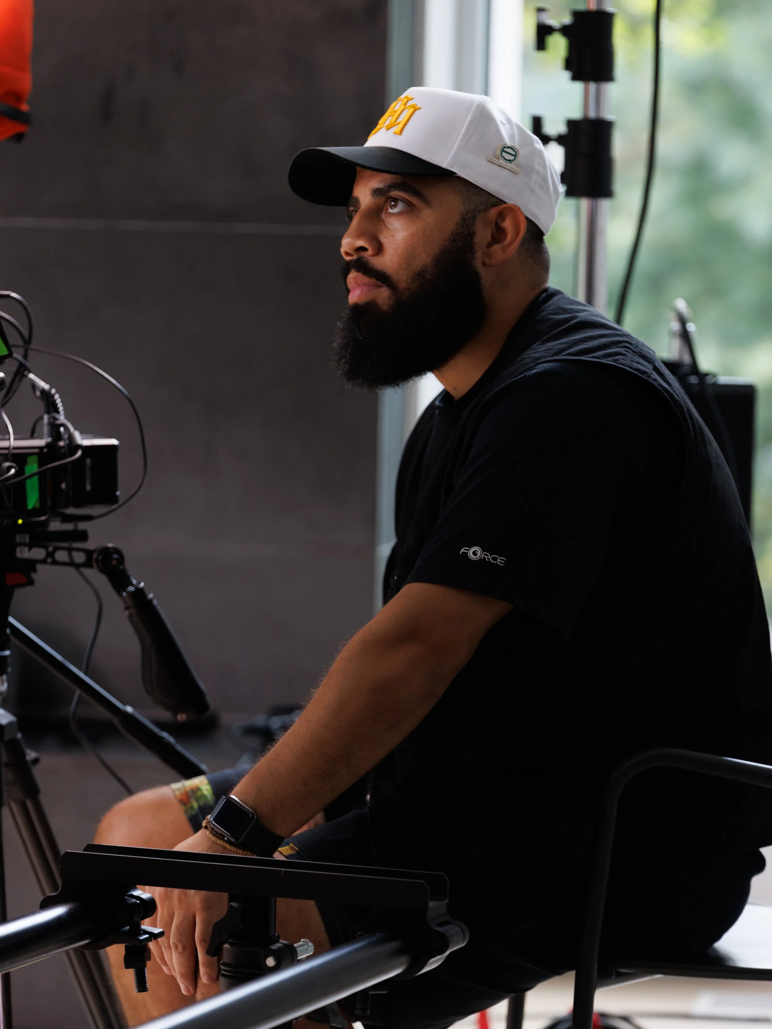 A man with a beard and wearing a white cap and black T-shirt sitting on a chair next to a camera setup, indoors near a window with greenery outside.