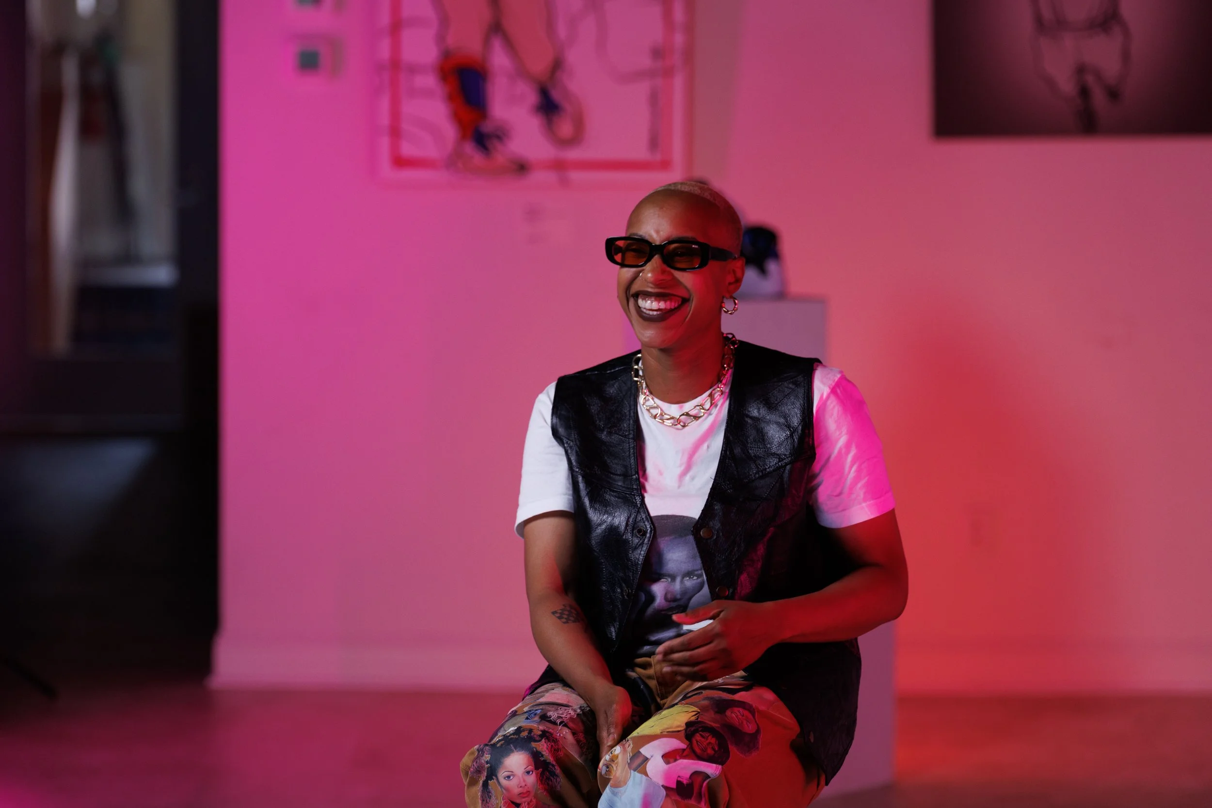 A person with a shaved head, glasses, and earrings is sitting in front of a pink background, smiling widely. They are wearing a graphic T-shirt with images of women and a black leather vest, along with colorful pants featuring portraits of women.