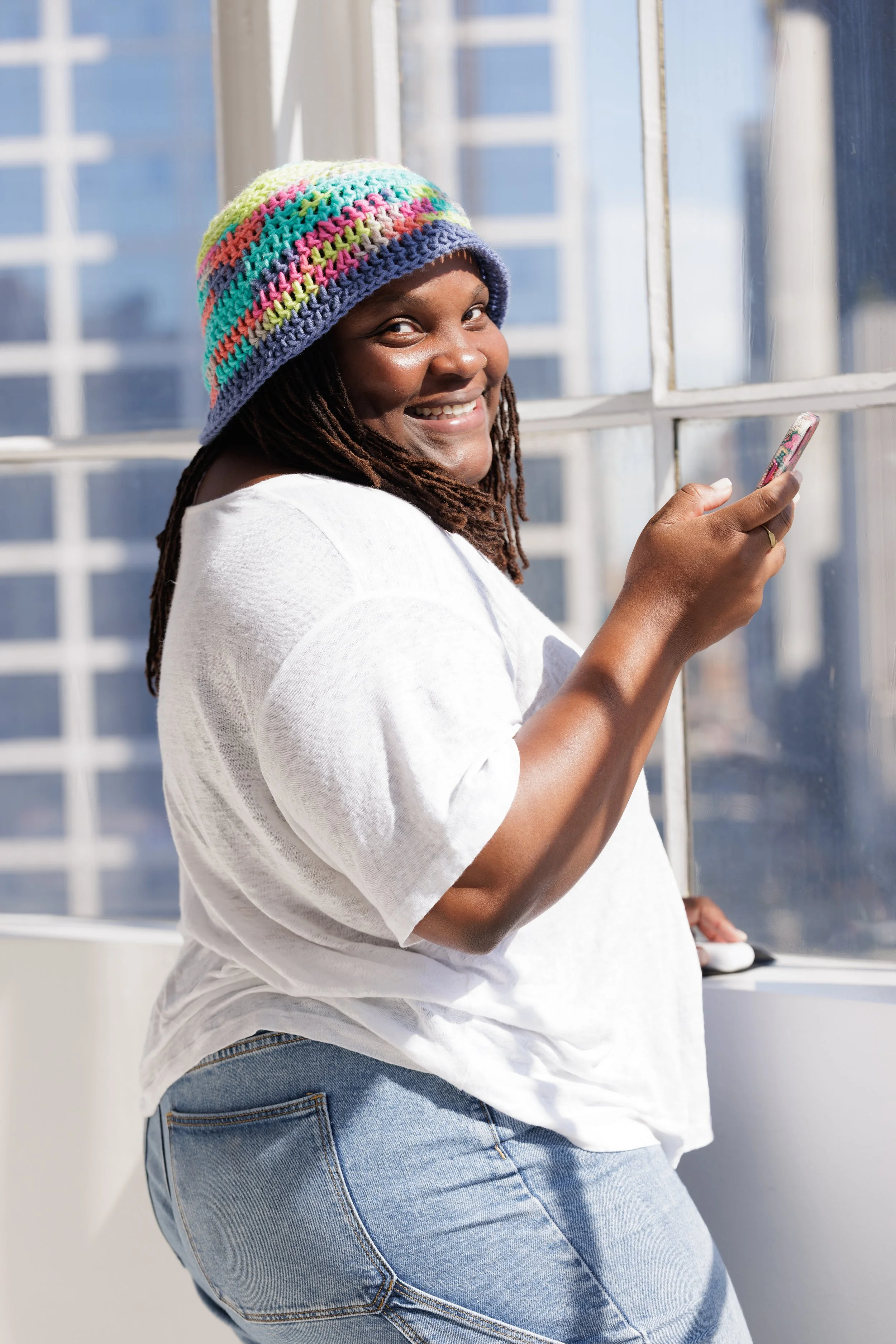 A woman in a white shirt and jeans, wearing a colorful knitted hat, smiling and looking at her phone while standing by a window with city buildings in the background.