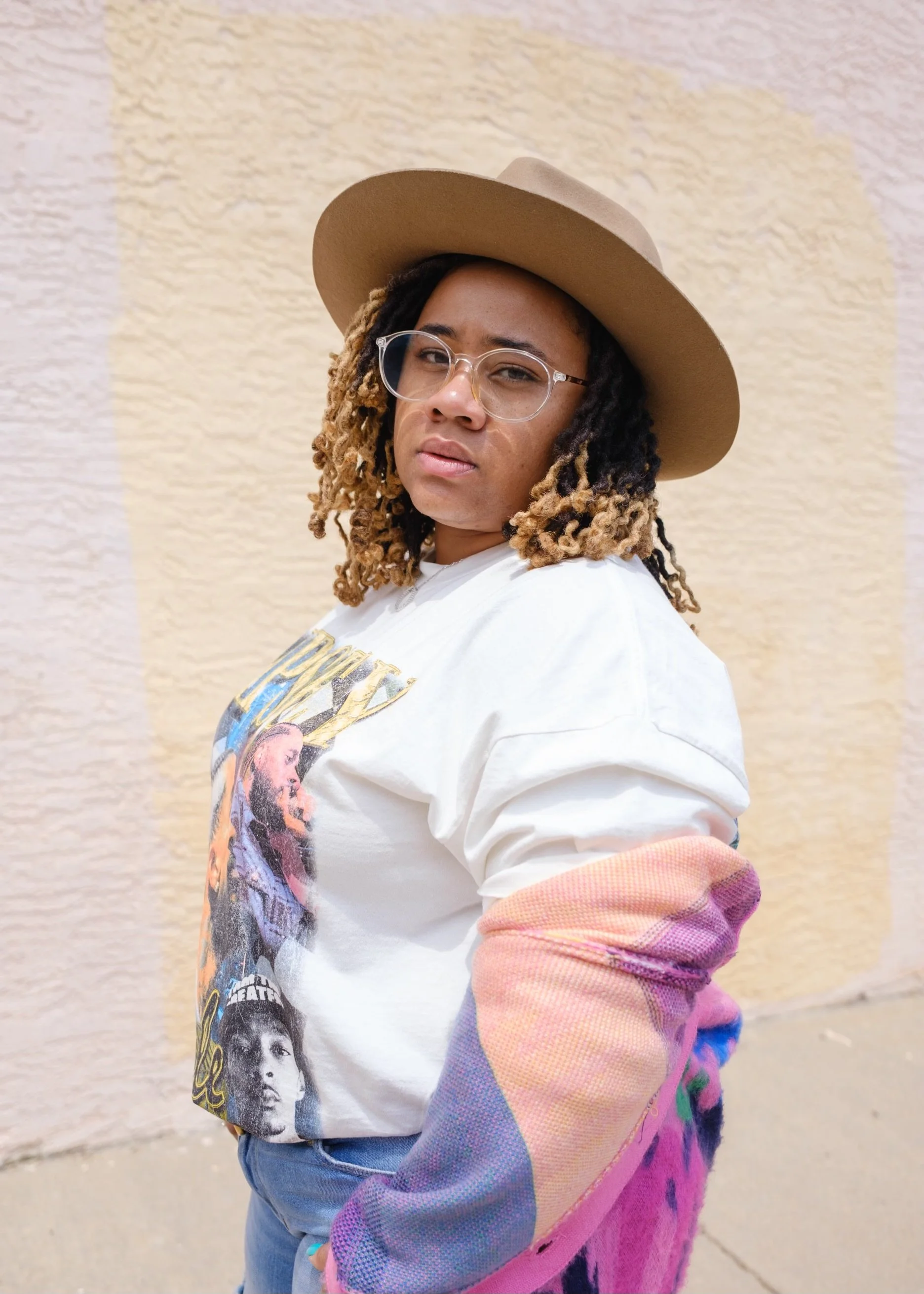 Woman with glasses, a wide-brimmed tan hat, dreadlocks, and a white graphic T-shirt standing in front of a textured beige wall.