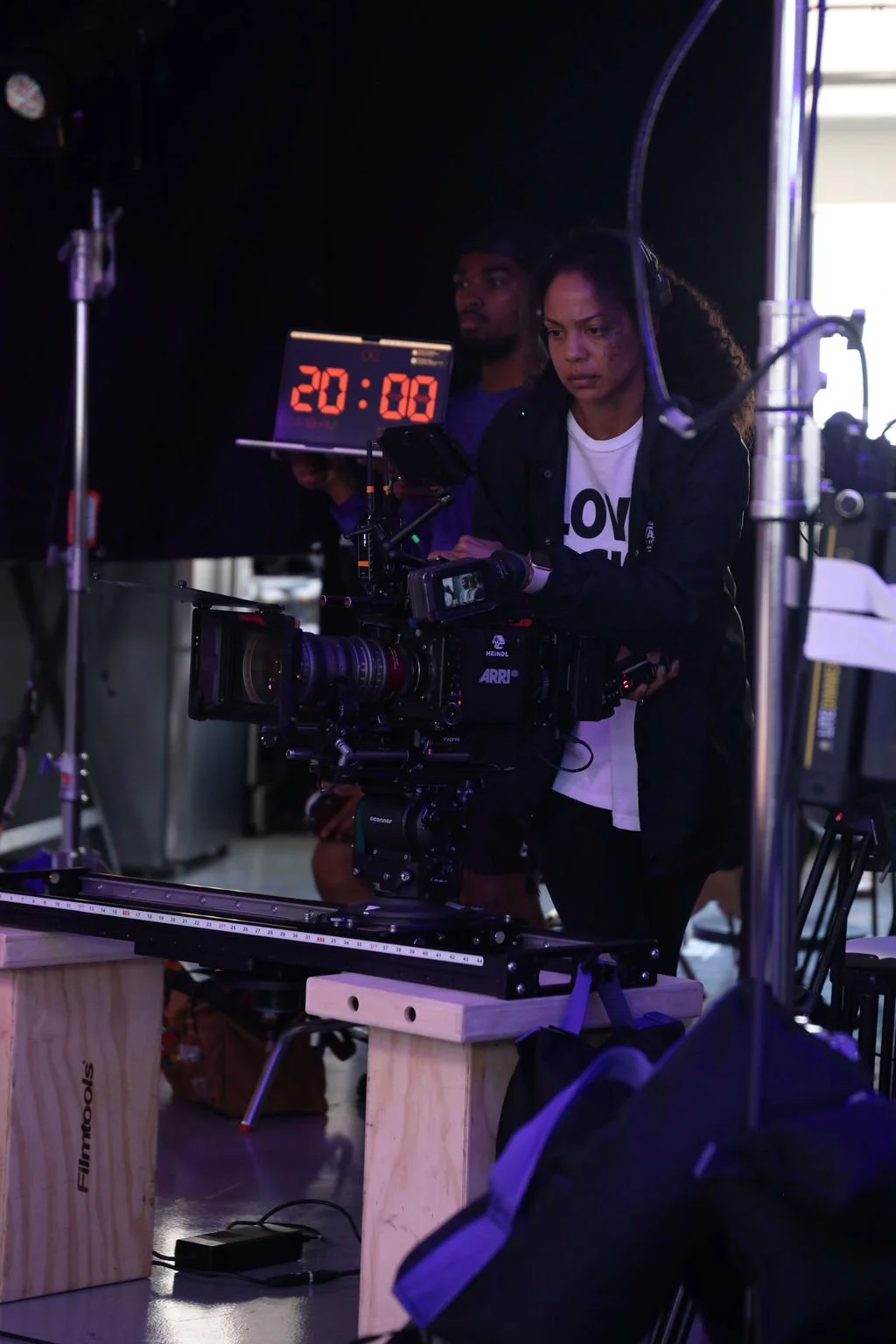 Two people working with a professional video camera setup in a dark studio environment with a digital countdown clock showing 20:08.