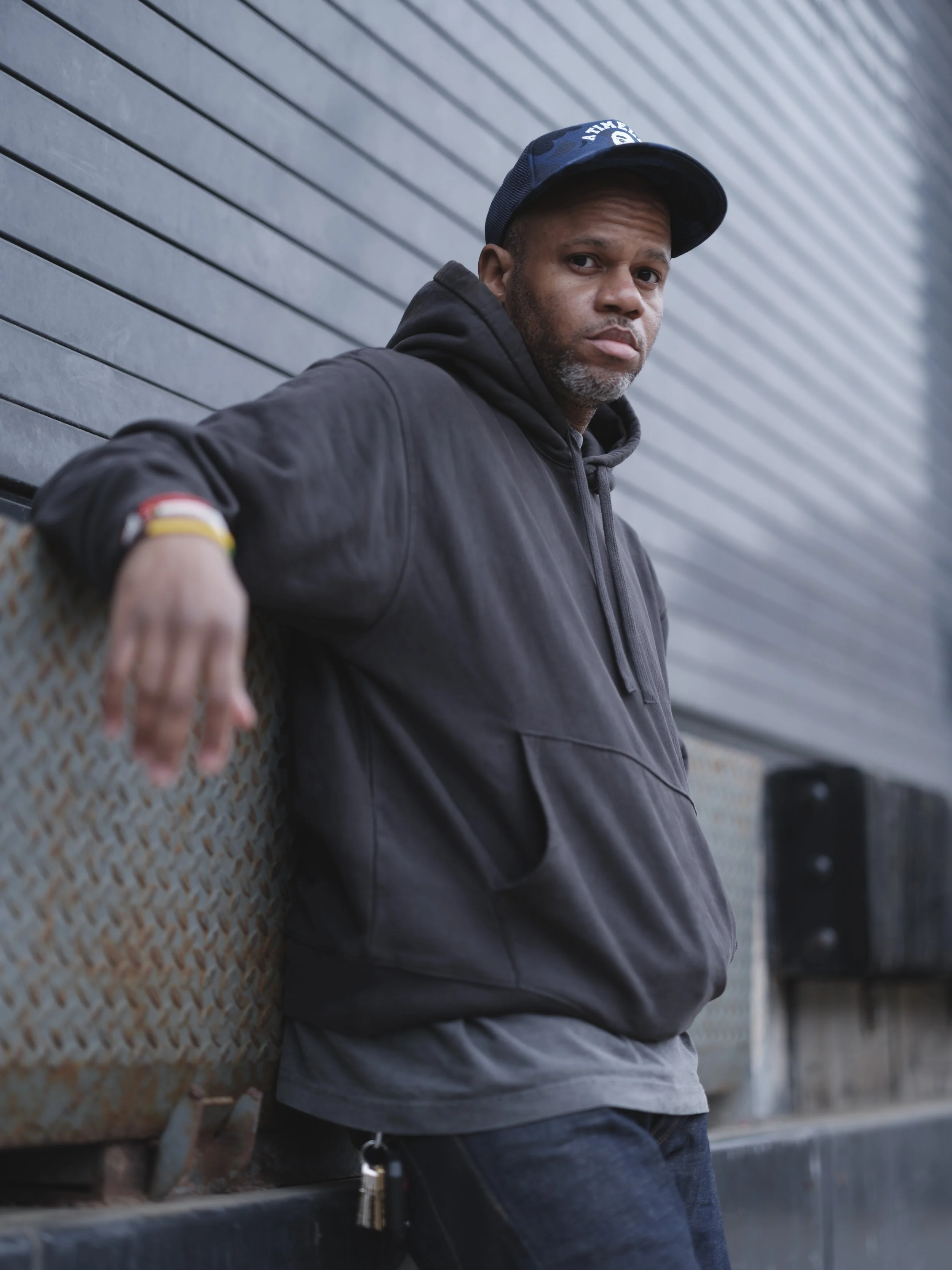 A man wearing a black hoodie and a blue cap leaning against a rusty metal surface with a serious expression, gray background, and industrial setting.