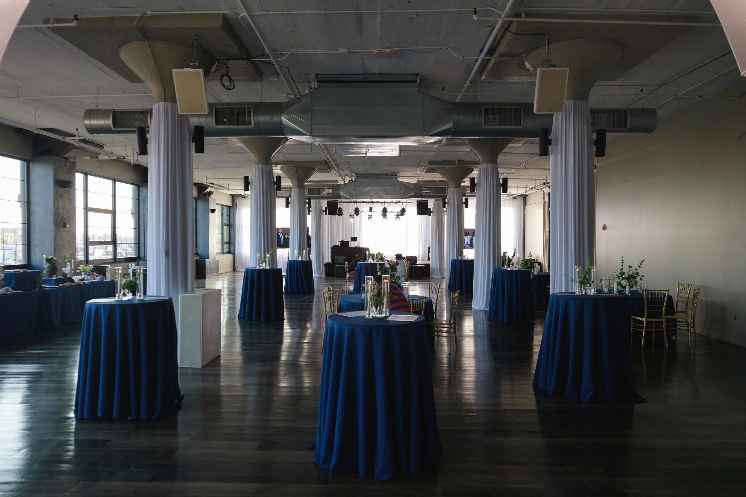 Wedding or event venue with round tables covered in blue tablecloths, white flower centerpieces, and gold chairs. Tall white curtains separate the space, with large windows allowing natural light. Ceiling has exposed ductwork and lighting equipment, creating an industrial-chic ambiance.
