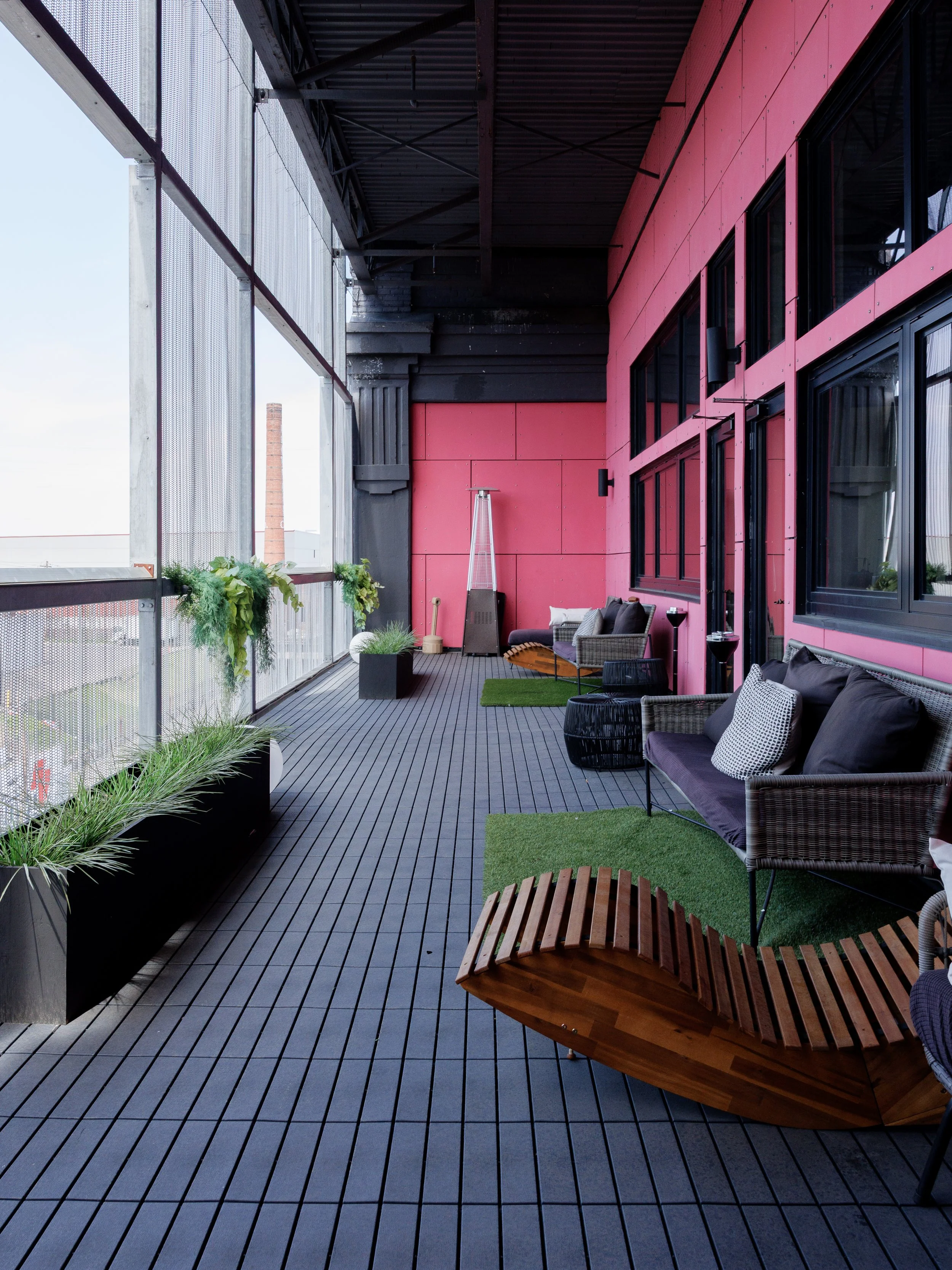 Empty outdoor balcony with tiled floor, pink wall with black window frames, and industrial elements in the background.