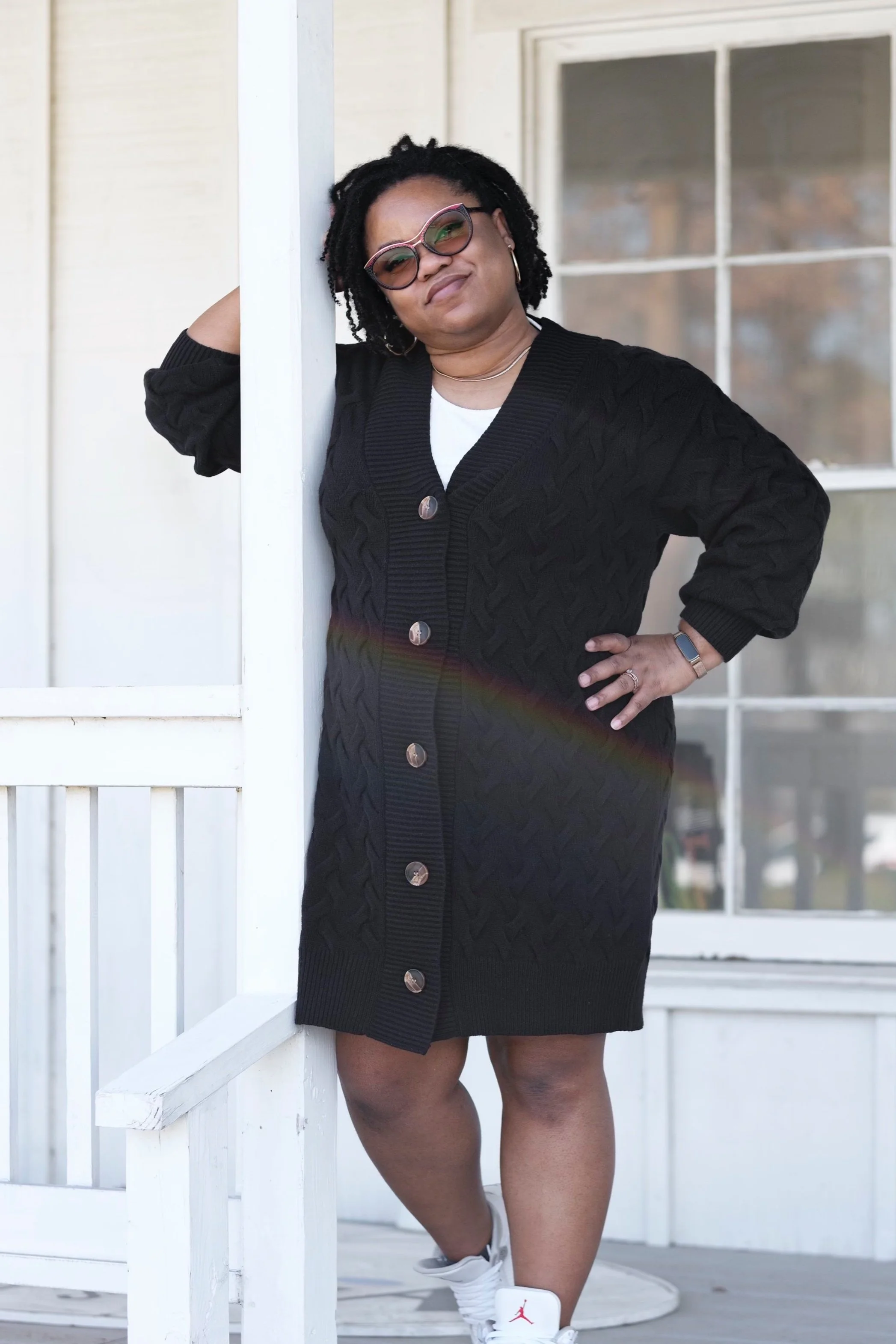 A woman with glasses and hoop earrings poses outside next to a white porch railing, wearing a black knitted cardigan with large buttons, white sneakers, and sunglasses, smiling with one hand on her hip.