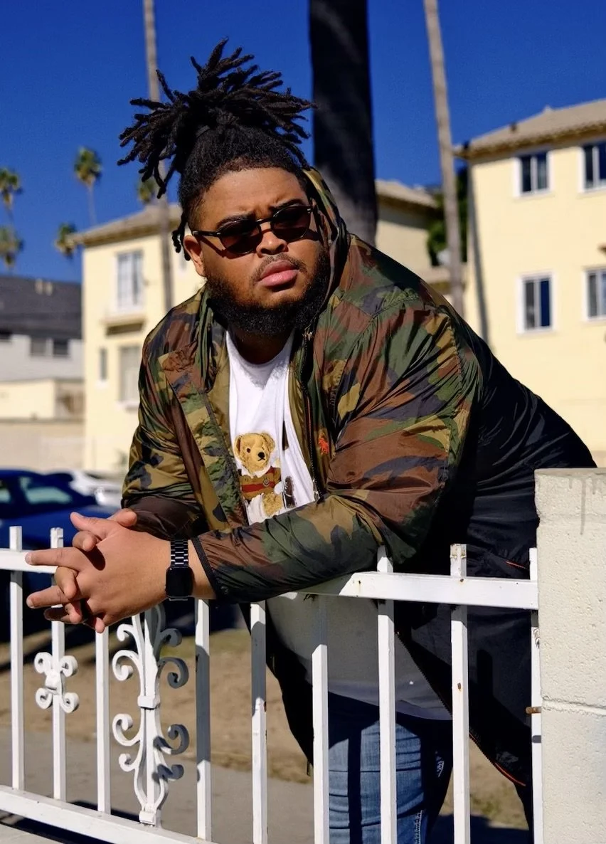 Man with dreadlocks and sunglasses leaning on a white fence, wearing a camouflage jacket over a white T-shirt with a teddy bear print, outdoors with residential buildings and palm trees in the background.