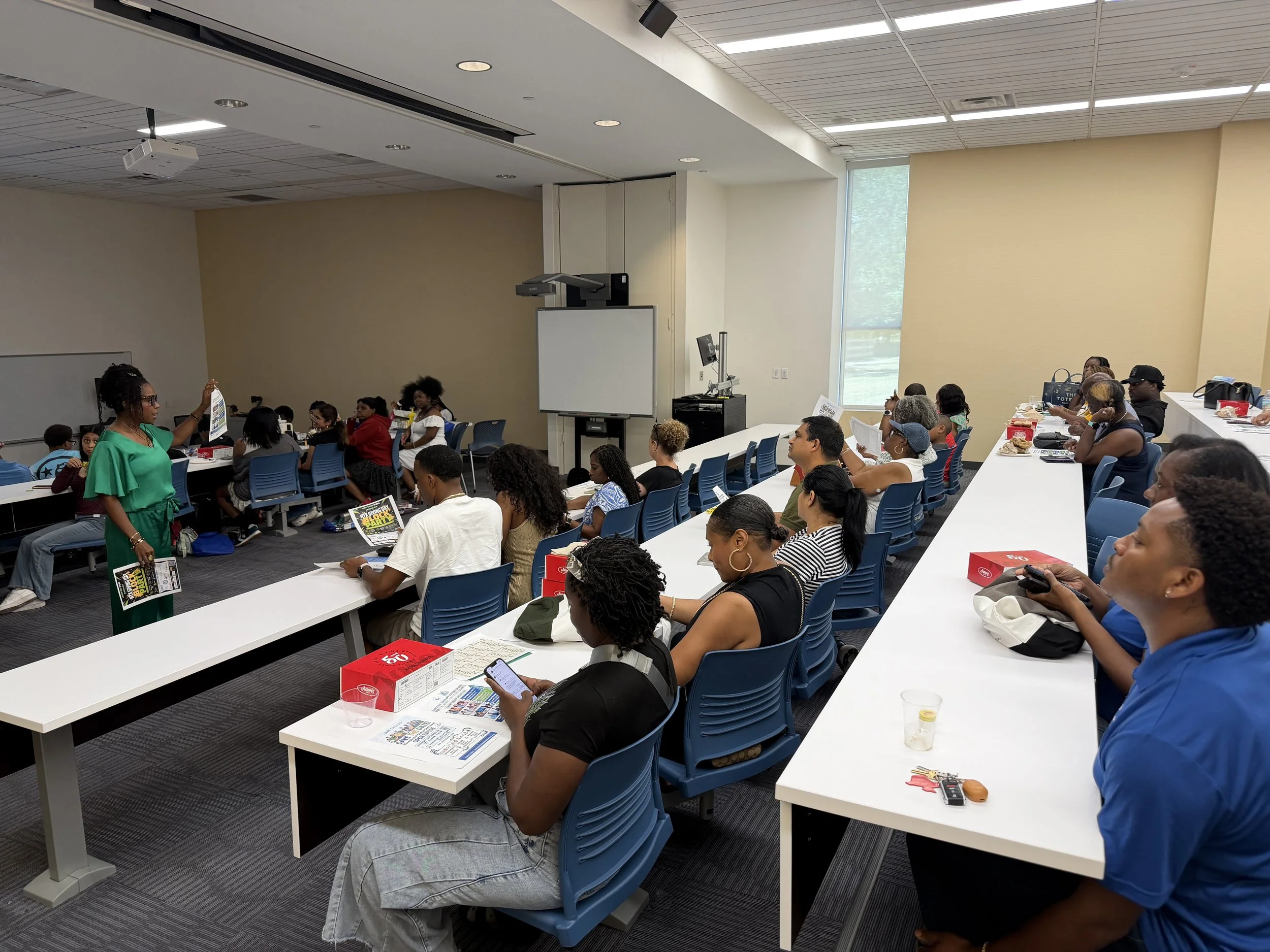 A diverse group of people attending a presentation in a classroom or conference room. They are seated at tables and listening to a woman in a green dress who is speaking and holding up a brochure. The room has a high ceiling, large windows, and a whiteboard at the front. There are boxes of snacks and papers on the tables.