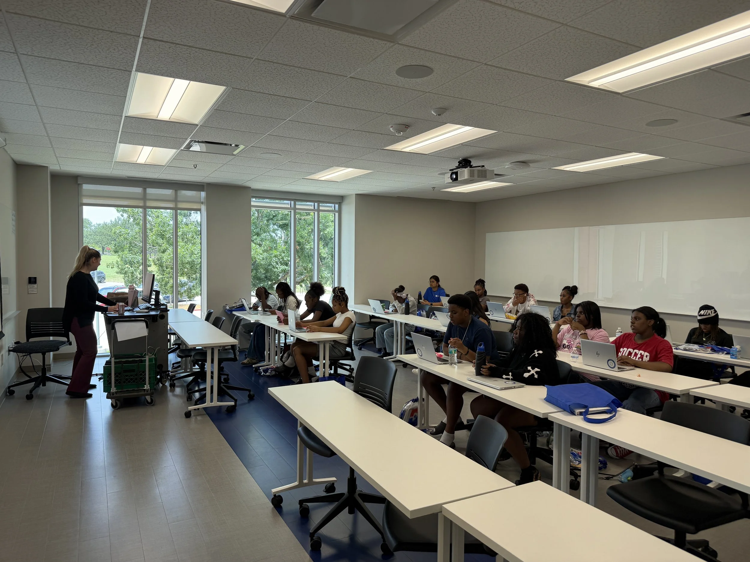 A classroom with students sitting at desks, a teacher at the front, large windows showing outside greenery, ceiling lights, and a projector.