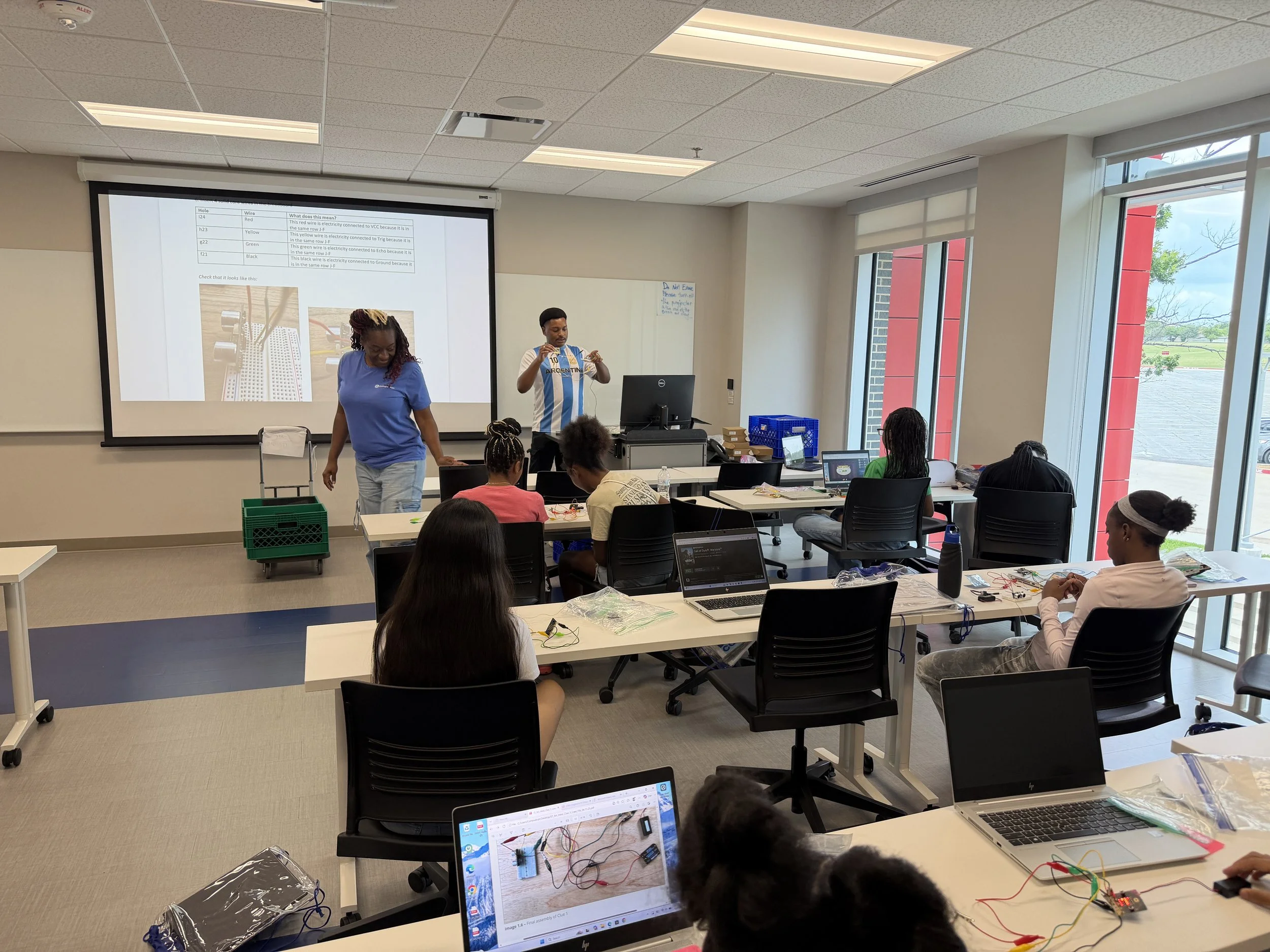 A classroom with students working on electronics projects, a teacher standing at the front, and a large screen displaying instructions and images of electronic components.