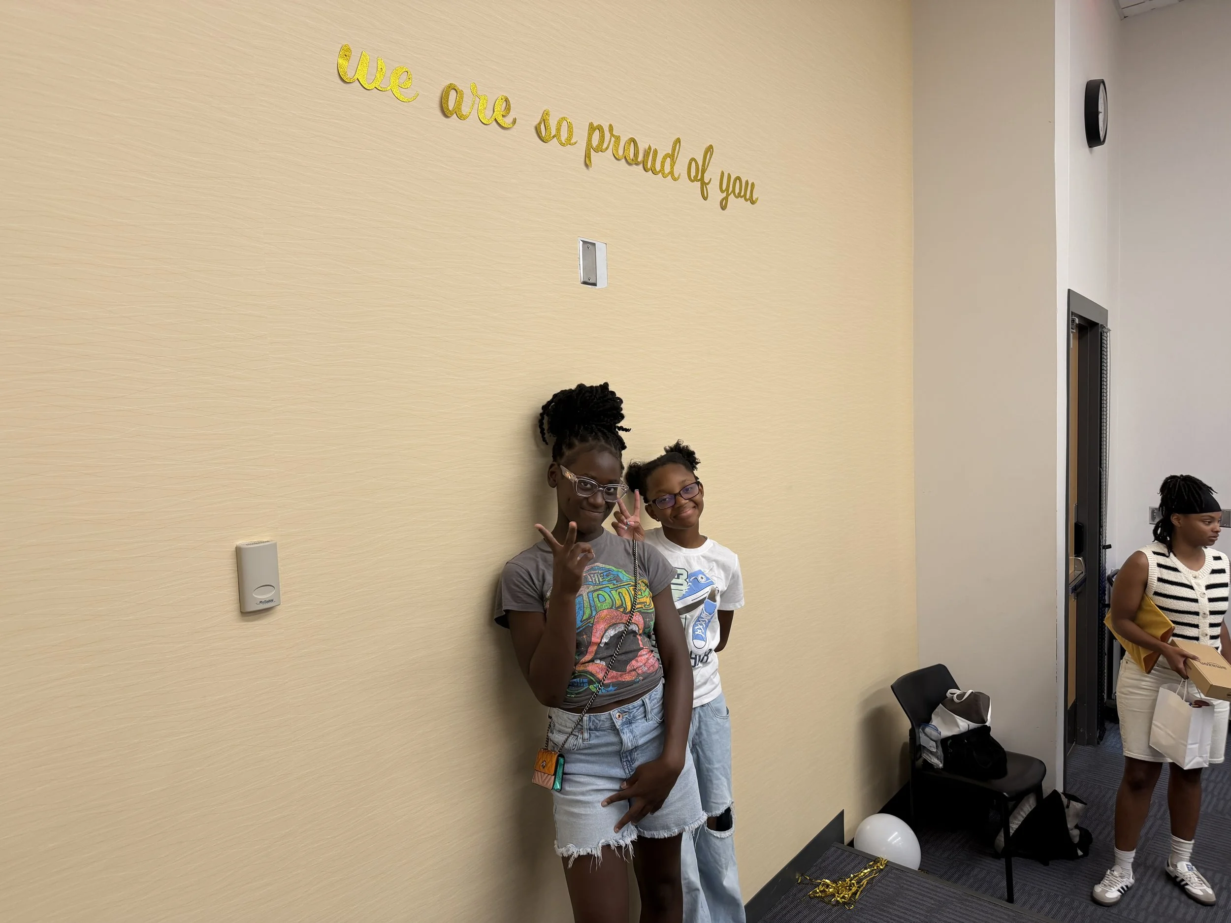 Two young girls standing and smiling against a beige wall that has a gold glitter sign reading 'we are so proud of you'. One girl is making a peace sign with her fingers.