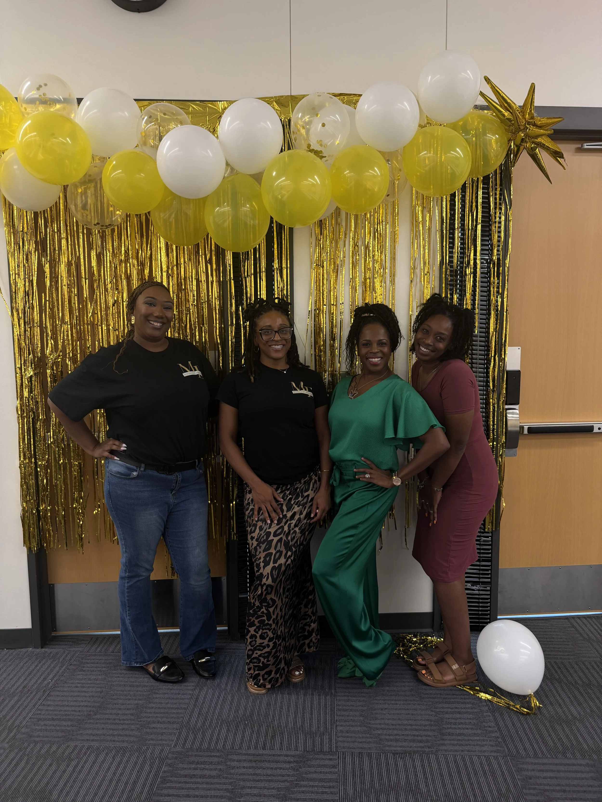 Four women standing in front of a gold and white balloon backdrop with gold and black streamers, smiling for the camera at a celebration event.