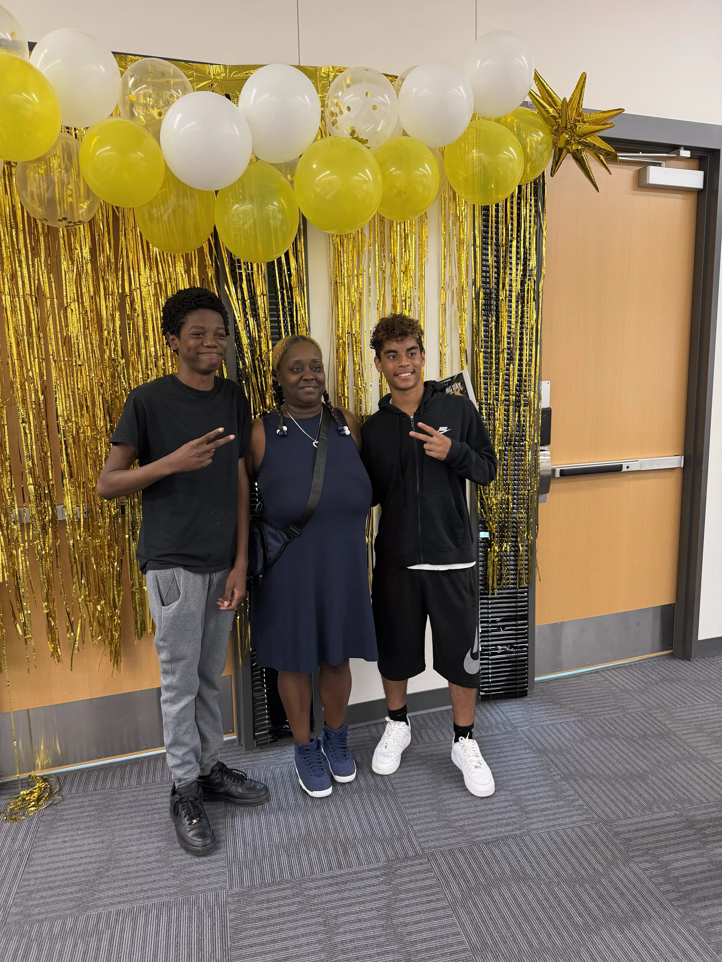 Three people standing in front of a gold balloon and streamers backdrop, making peace signs with their hands.