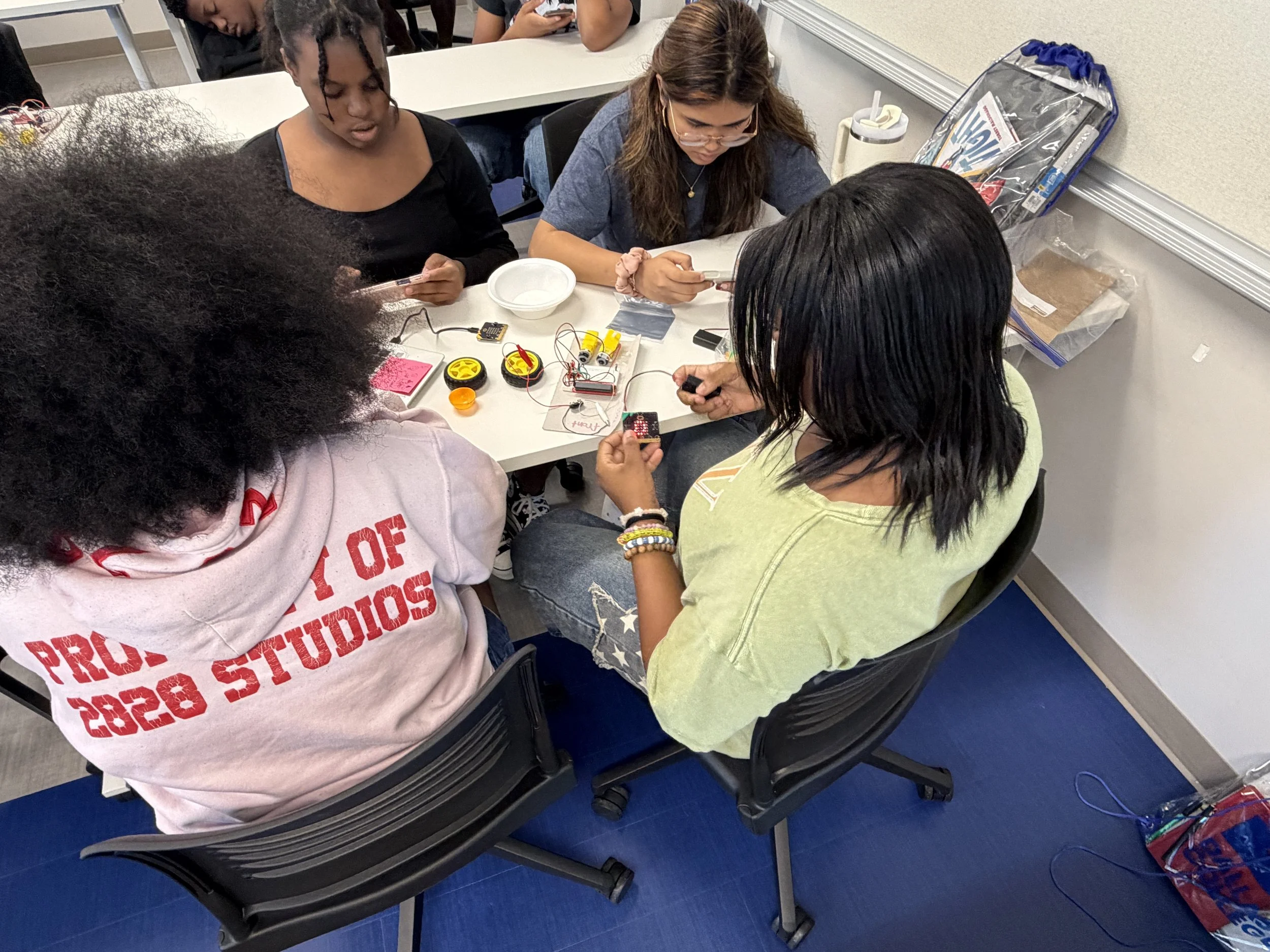 A group of diverse students sitting around a table working on a robotics project with electronic components, wires, and devices, in a classroom setting.