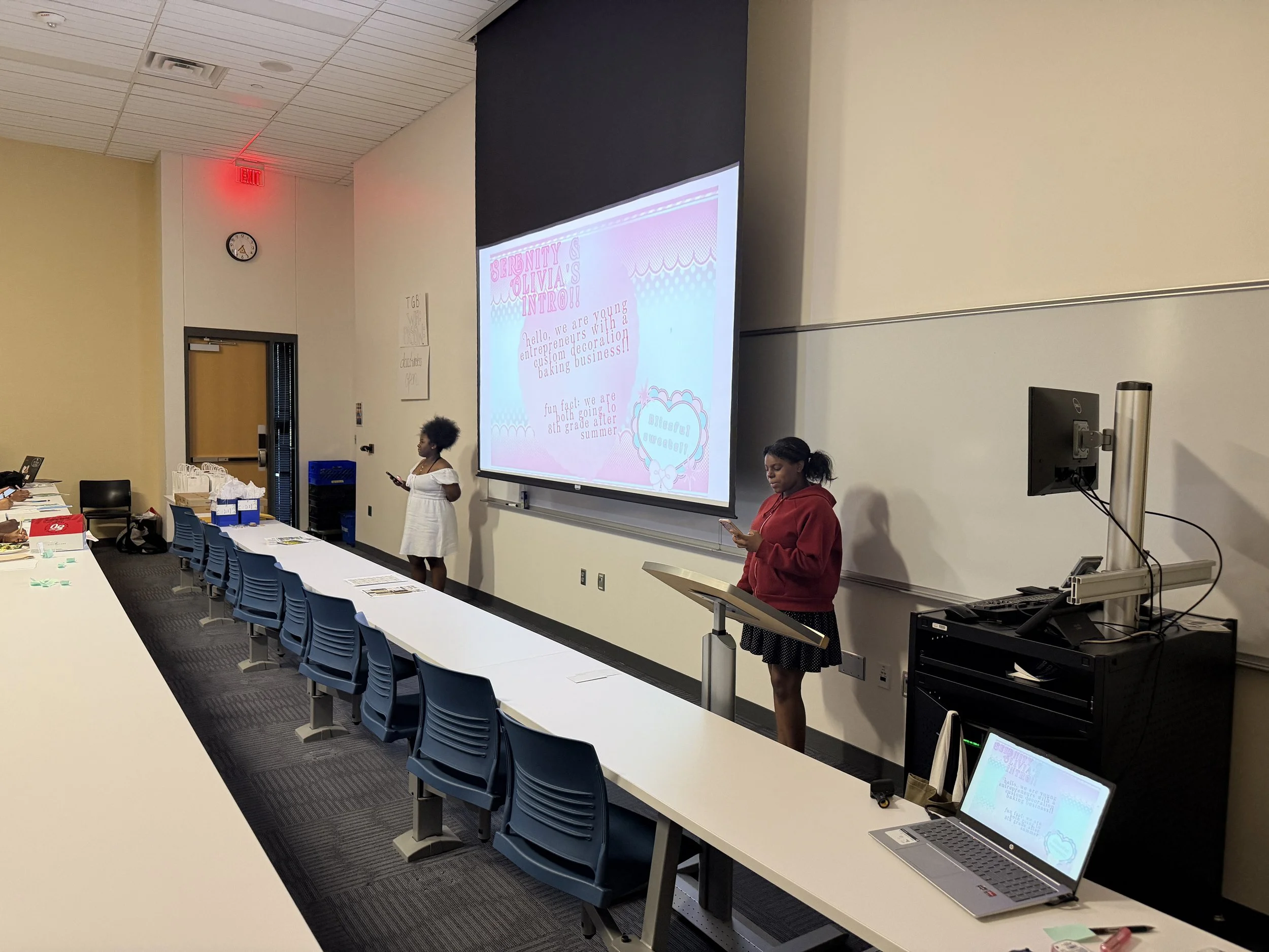 A classroom with two women standing at the front near a large screen displaying a presentation titled 'Birthdays & Olivia's Intro!'. One woman is wearing a white dress, holding a phone, and the other is in a red hoodie, also holding a phone. There are tables with papers and supplies, and a few people seated.