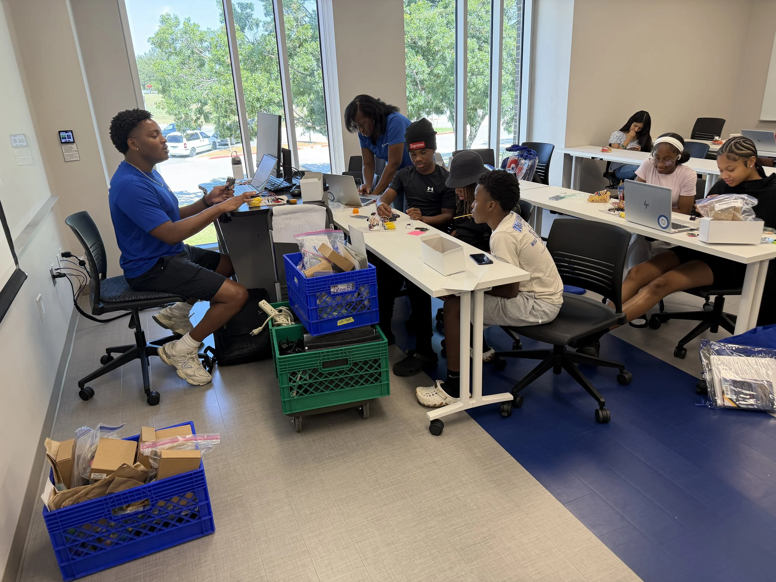 A classroom with students working at tables, some using laptops and electronic components, and a teacher assisting students. There are large windows showing trees and parked cars outside.