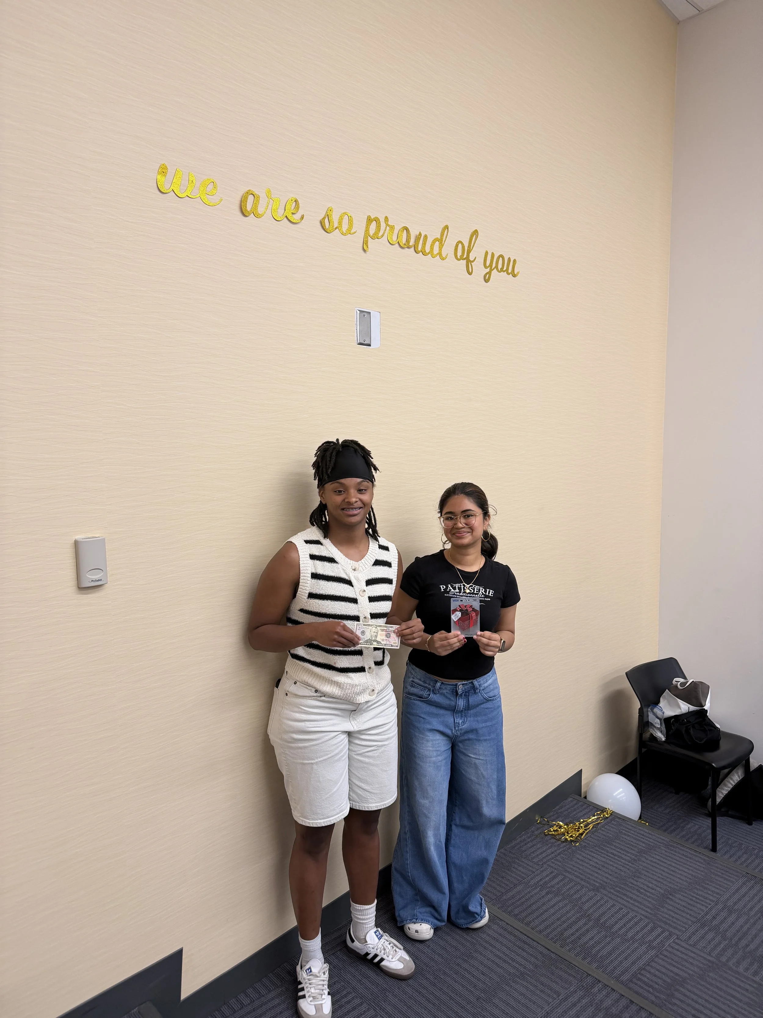 Two women standing against a beige wall, holding cash and a card, with a sign above that says, 'we are so proud of you' in gold lettering.