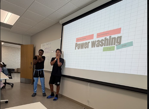 Two students standing in front of a classroom presentation slide titled 'Power washing.' The slide has colored tags with text and numbers, and a whiteboard is visible in the background.