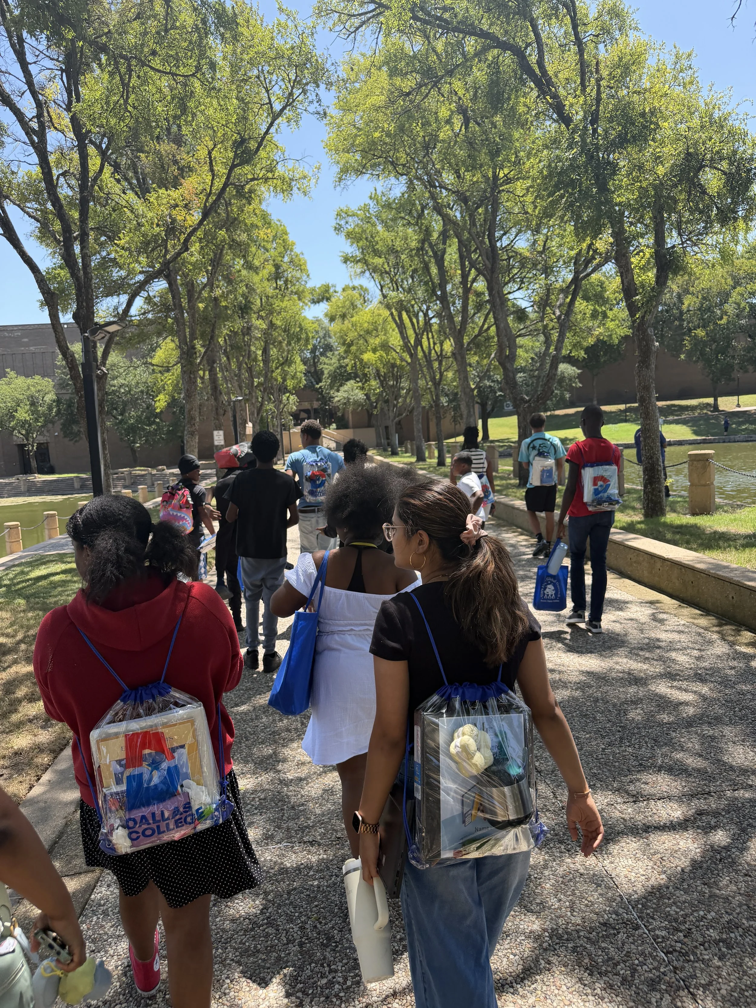 A group of people walking outdoors along a paved path with trees on both sides, some carrying backpacks and tote bags, with a building and grassy area in the background on a sunny day.