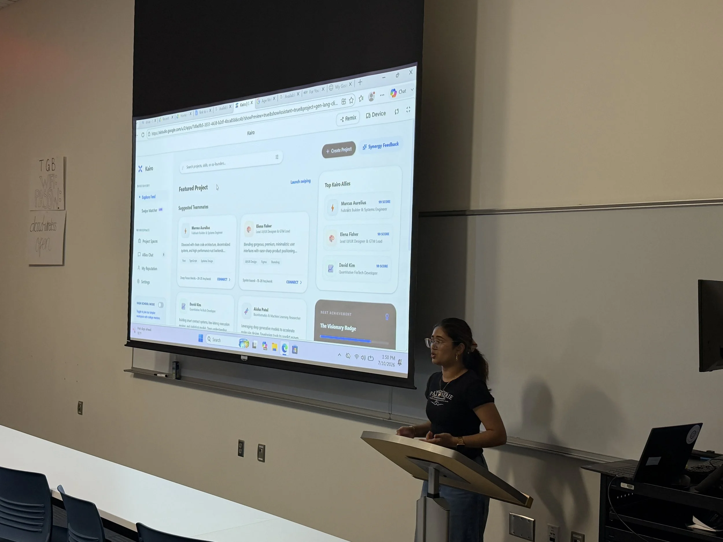 A woman standing at a lectern in front of a classroom, giving a presentation. A large screen behind her displays a computer desktop with a project management web app open. The classroom has white walls, a row of black chairs, and some posters on the wall.