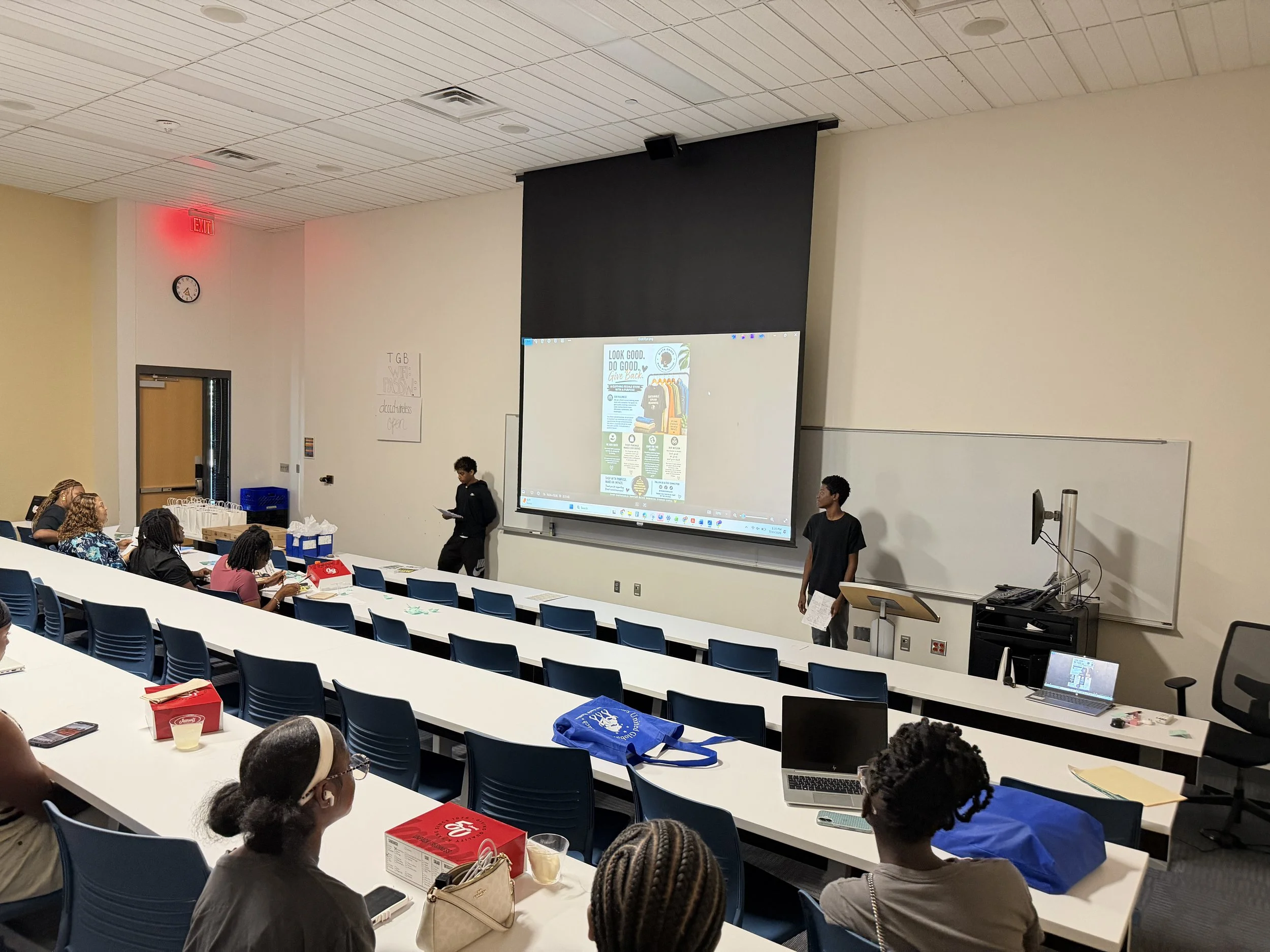 People sitting in a classroom with a large projection screen at the front, and two individuals standing near it, one reading from paper, in an educational setting.