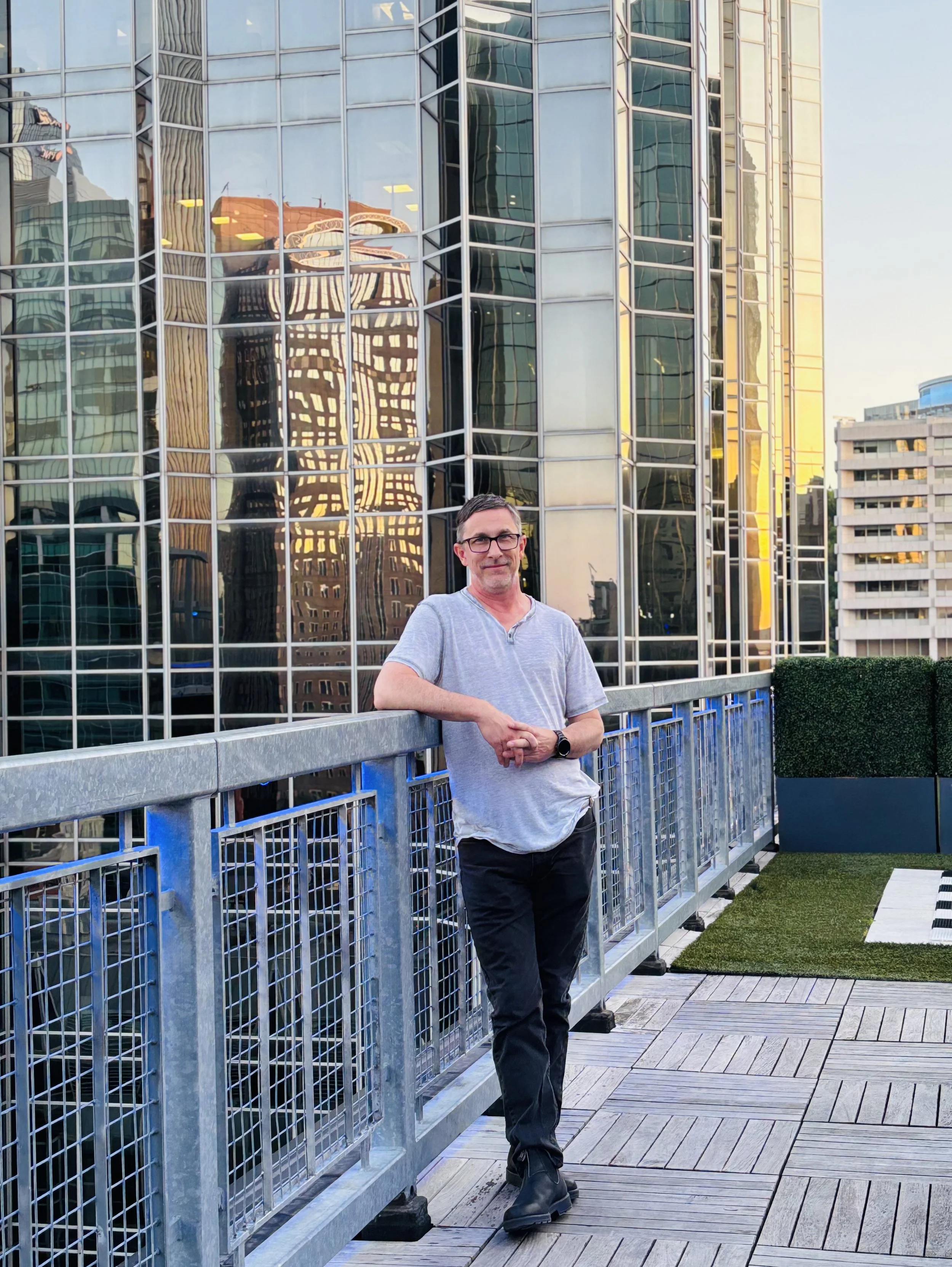 A man leaning on a metal railing on a rooftop terrace with a cityscape of curved glass skyscrapers reflecting nearby buildings in the background during sunset.